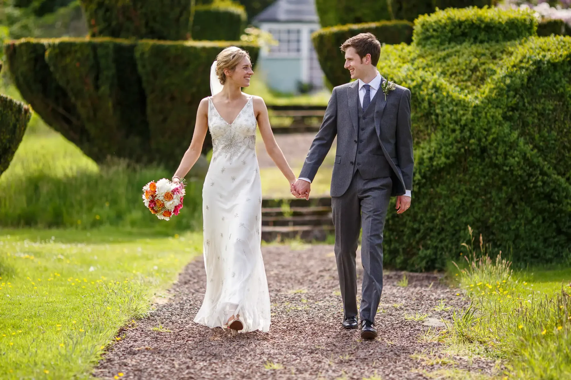 A bride in a white dress and a groom in a grey suit walk hand in hand on a garden path, smiling at each other. The bride holds a bouquet of flowers.