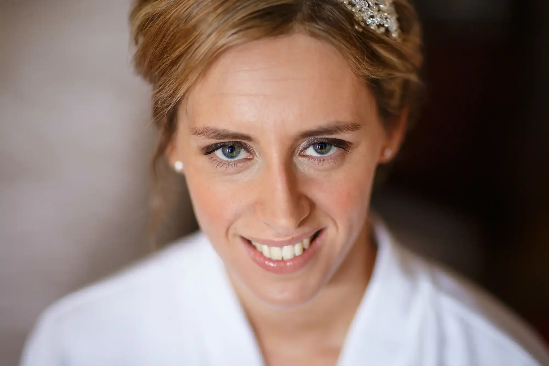 Close-up of a smiling woman with an updo hairstyle, wearing a white robe and a decorative hair accessory, with a soft focus background.