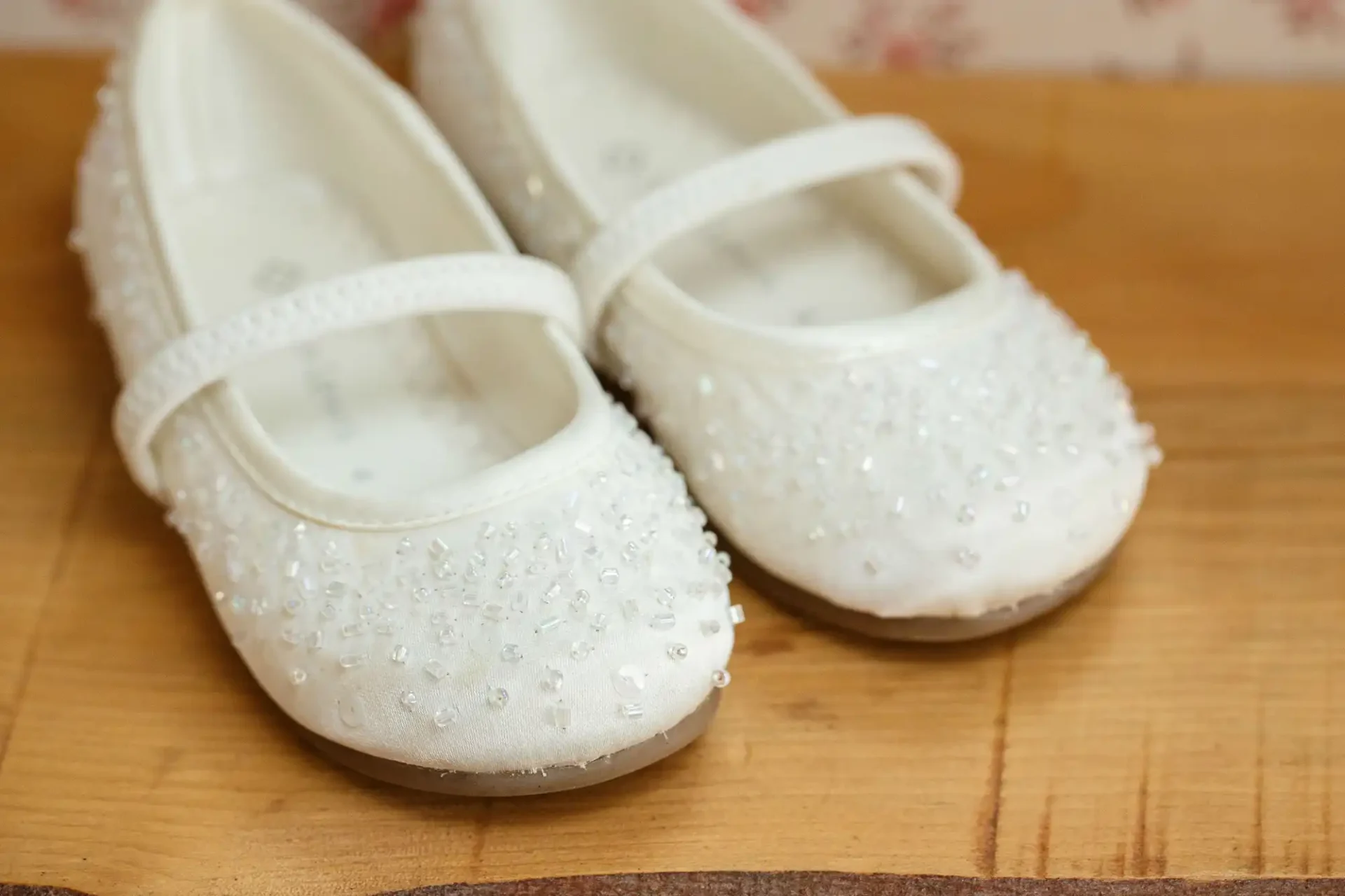 A pair of white, beaded children's ballet flats on a wooden surface.