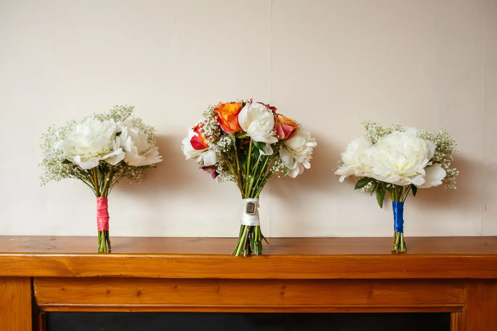 Three bridal bouquets with white and orange flowers wrapped in colorful ribbons, displayed on a wooden surface against a plain wall.