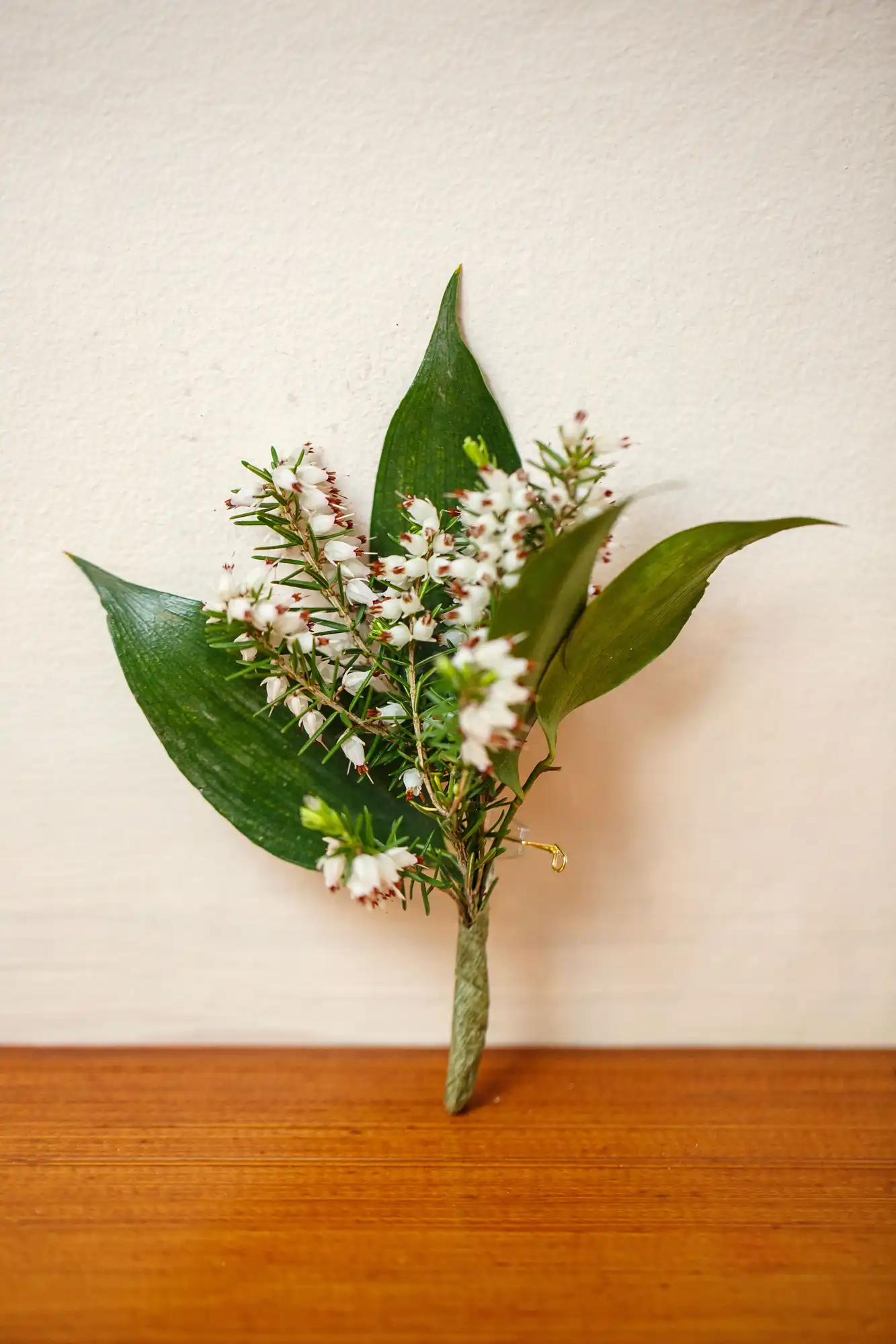Small bouquet of white bell-shaped flowers and green leaves, wrapped in a leaf, standing on a wooden surface against a white wall.