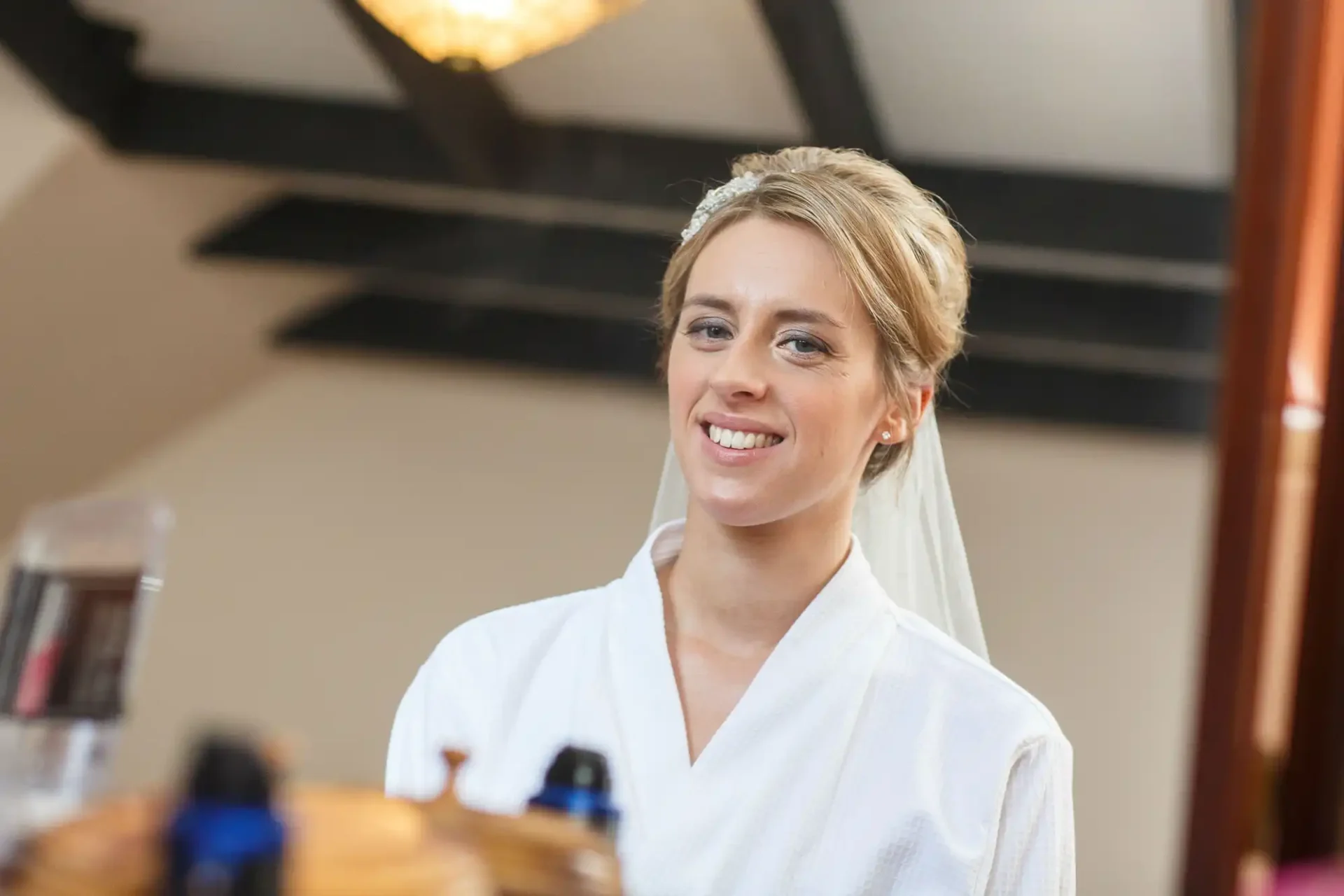 A smiling bride with short hair in a white robe and veil indoors, looking away from the camera.