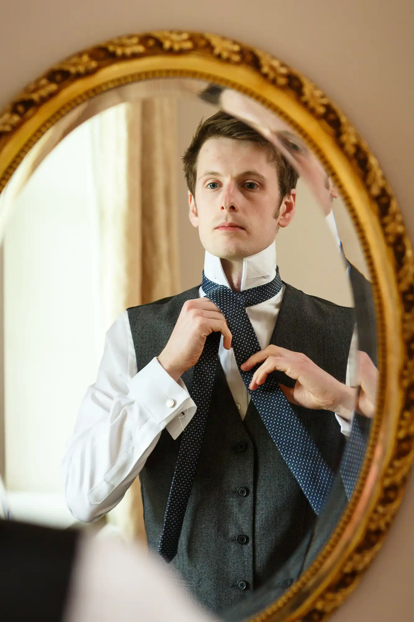 A man in a vest and dress shirt adjusts his tie while looking at himself in an ornate oval mirror.