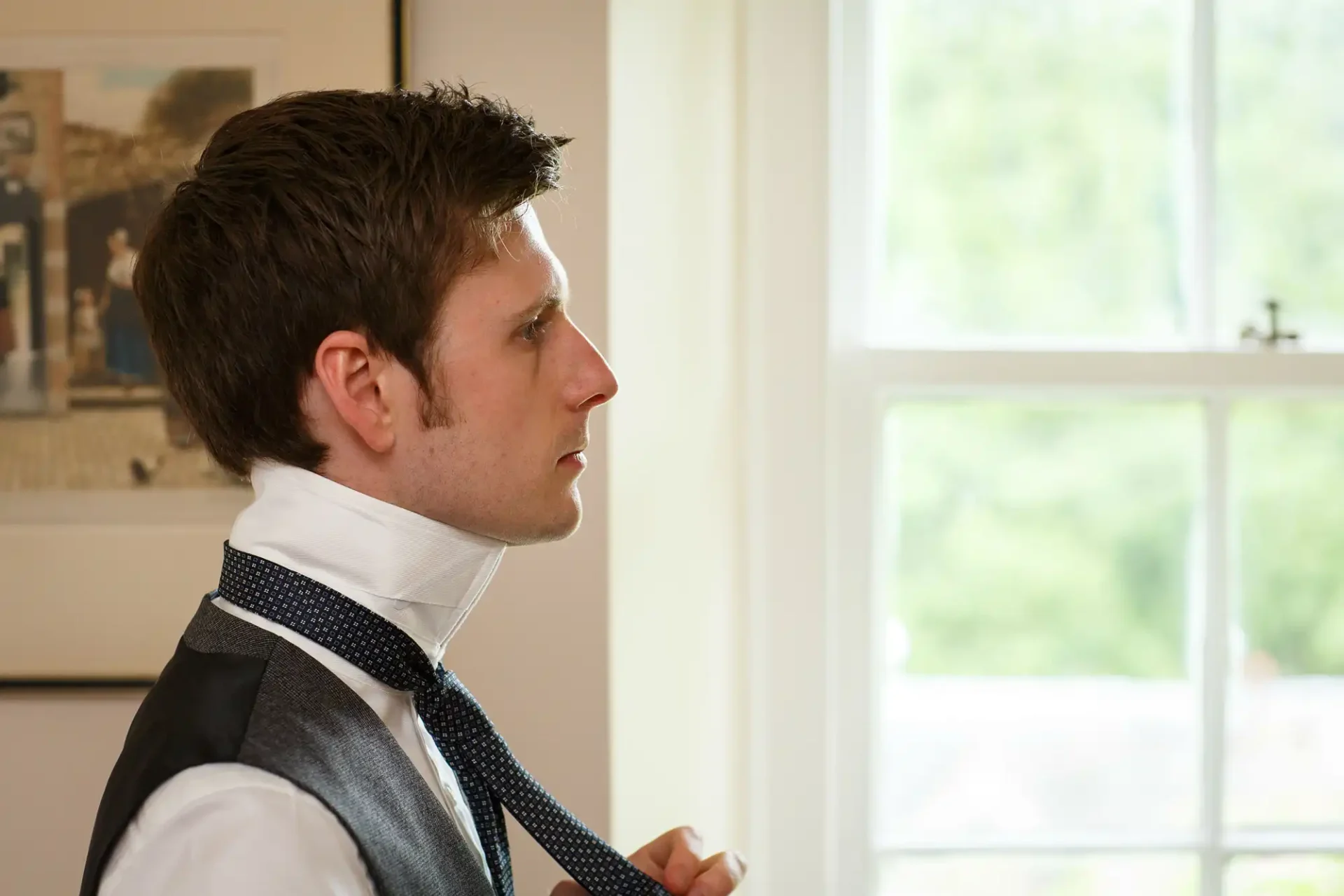 A man in a vest and tie adjusting his tie, looking off to the side with a focused expression, indoors with a window in the background.