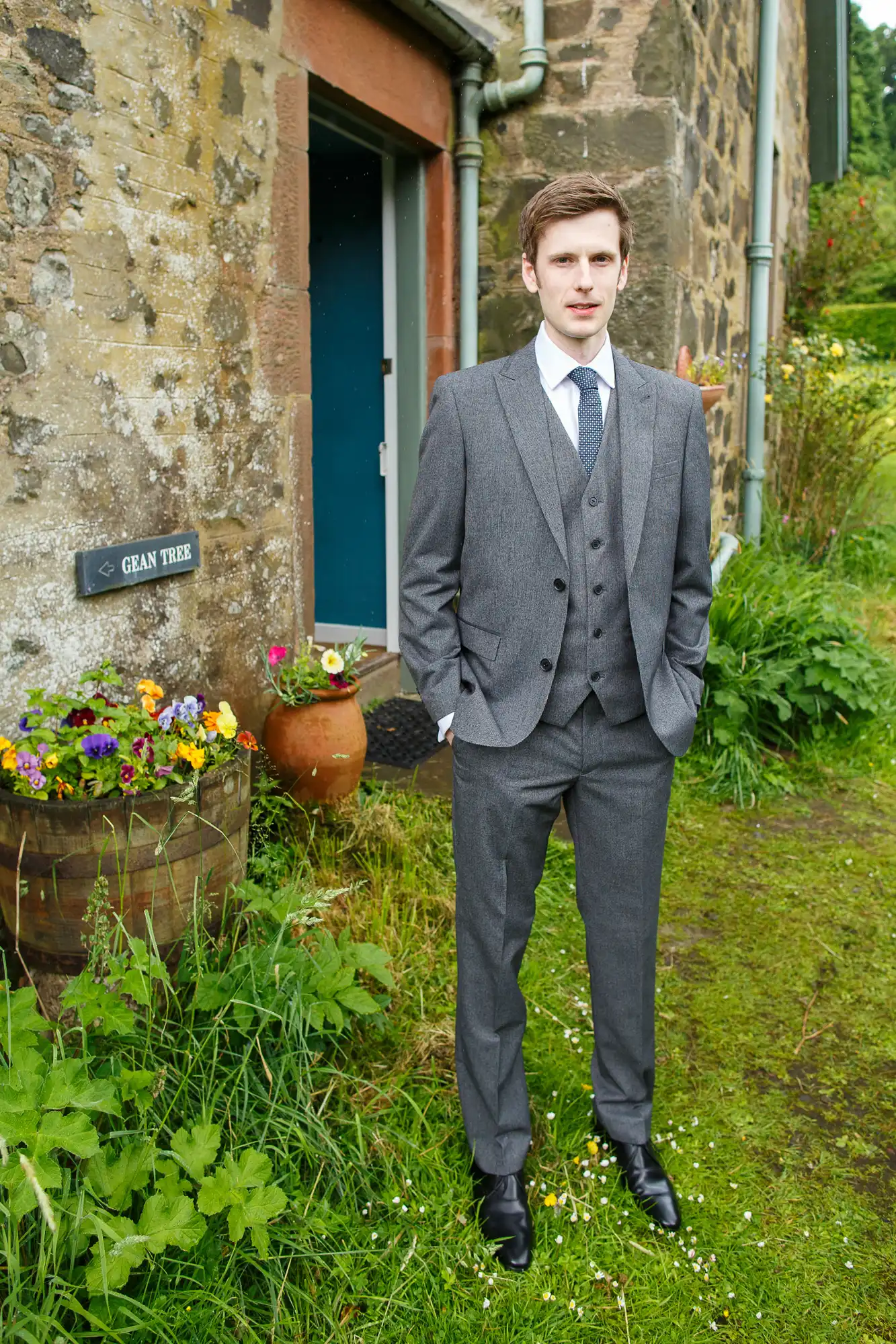 A young man in a formal gray suit and tie standing in front of a rustic stone building with a sign that reads "gran té" and surrounded by greenery and flowers.