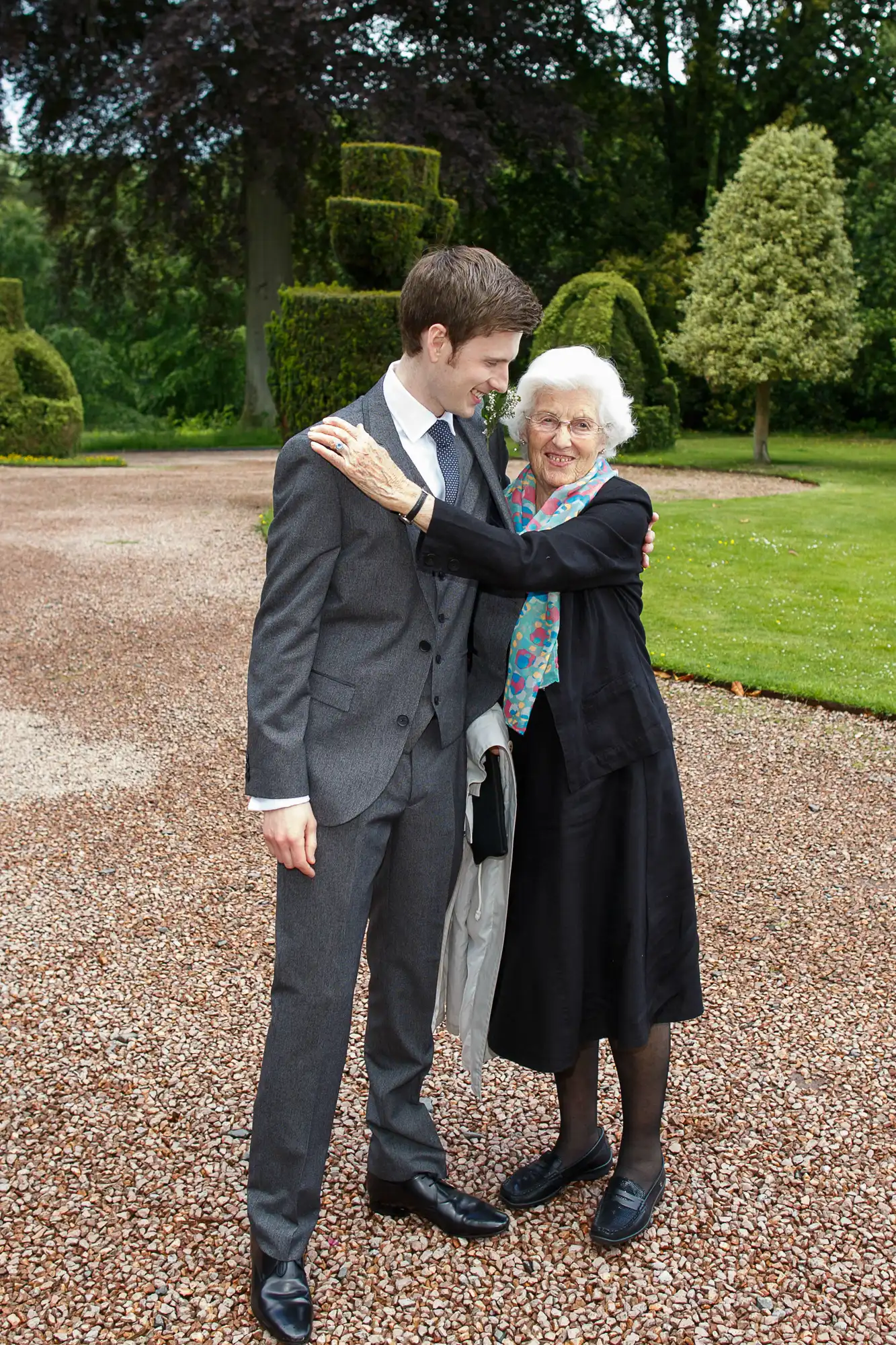 A young man in a suit and an elderly woman in a black dress and colorful scarf embrace, smiling, in a garden with topiary trees.