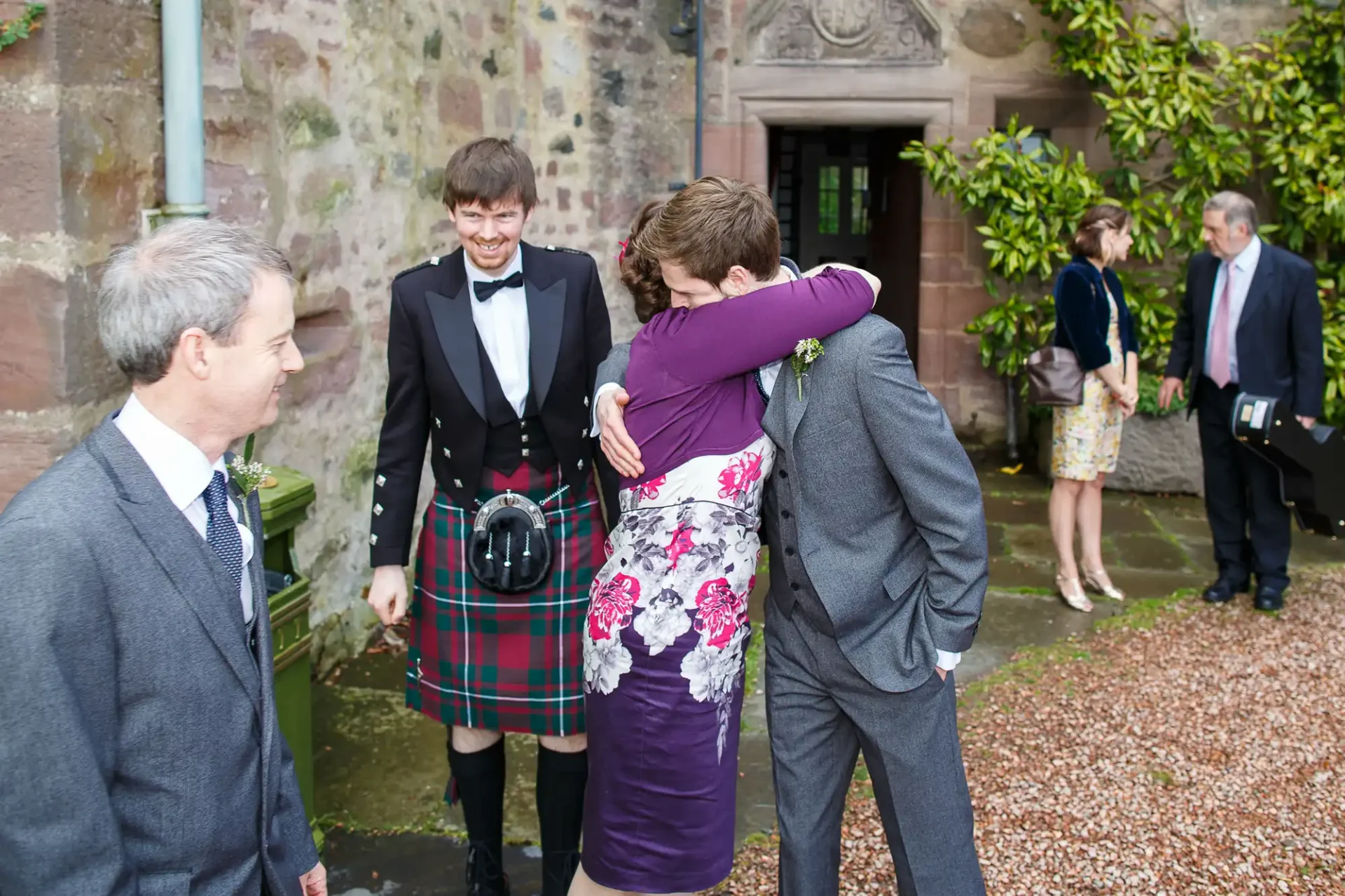Two men embrace warmly at an event, one in a tartan kilt, while another man and two women observe, all dressed in formal wear, outside a stone building with vines.