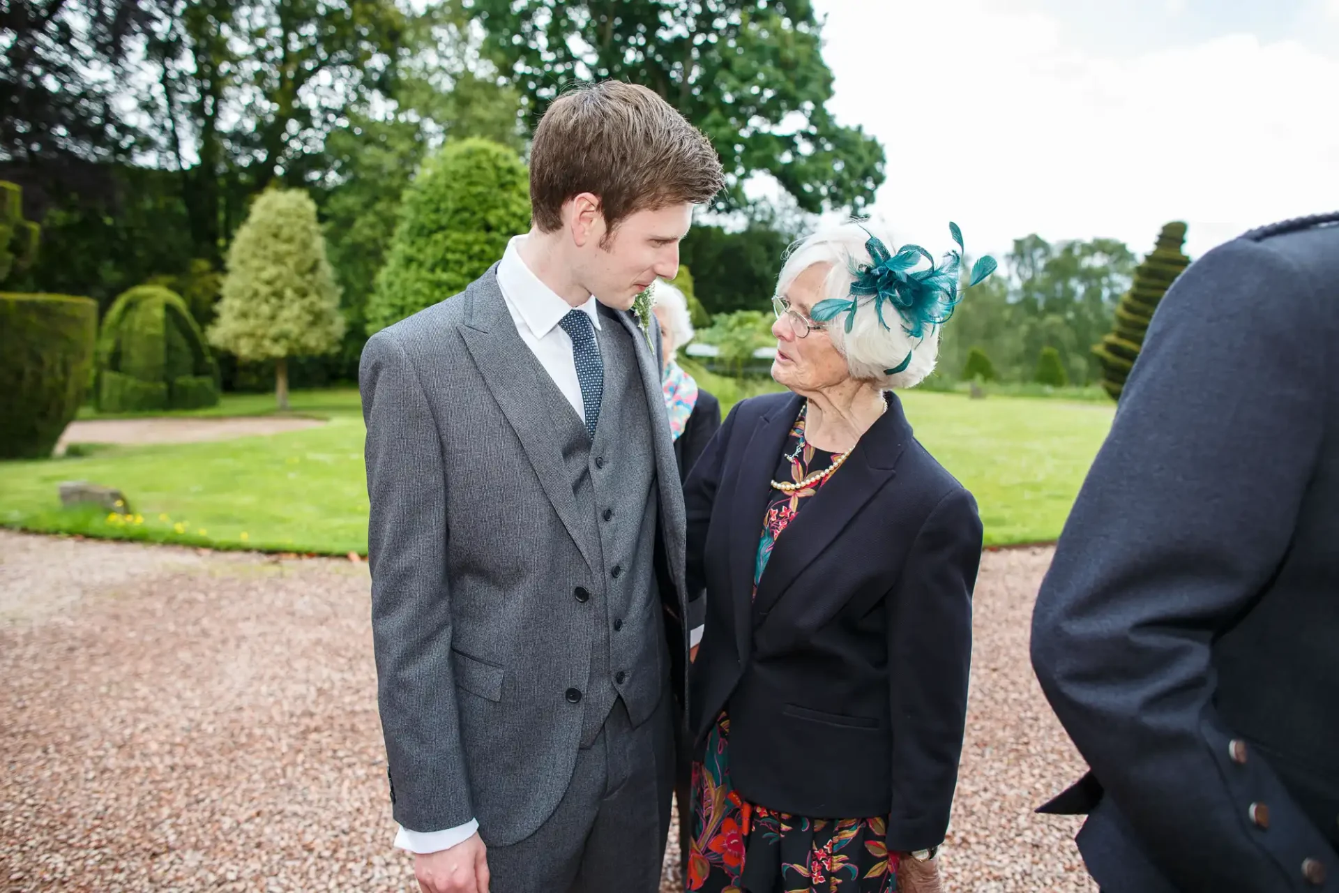 A young man in a grey suit looks affectionately at an elderly woman with a blue fascinator and floral dress at a garden event.
