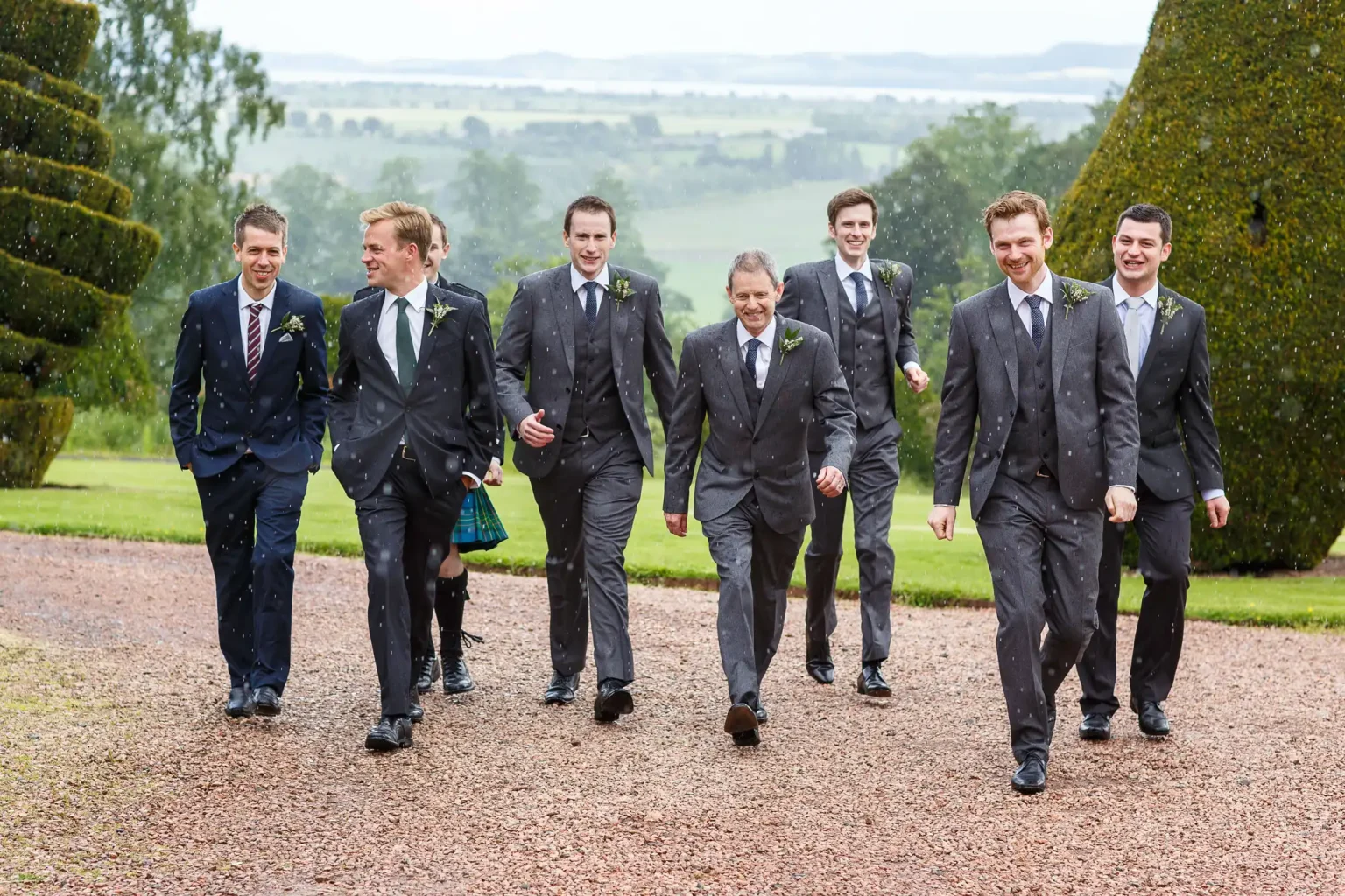 Seven men in suits walk outside on a gravel path, with a grassy landscape and overcast sky in the background. The two leftmost individuals wear ties with stripes, while the others have solid-coloured ties at Fingask Castle, Perth and Kinross.