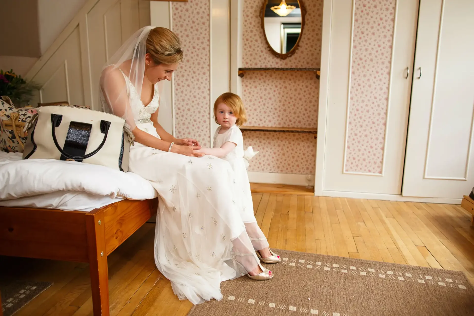 A bride in a white dress sitting and holding hands with a young child in a white dress in a warmly lit room.