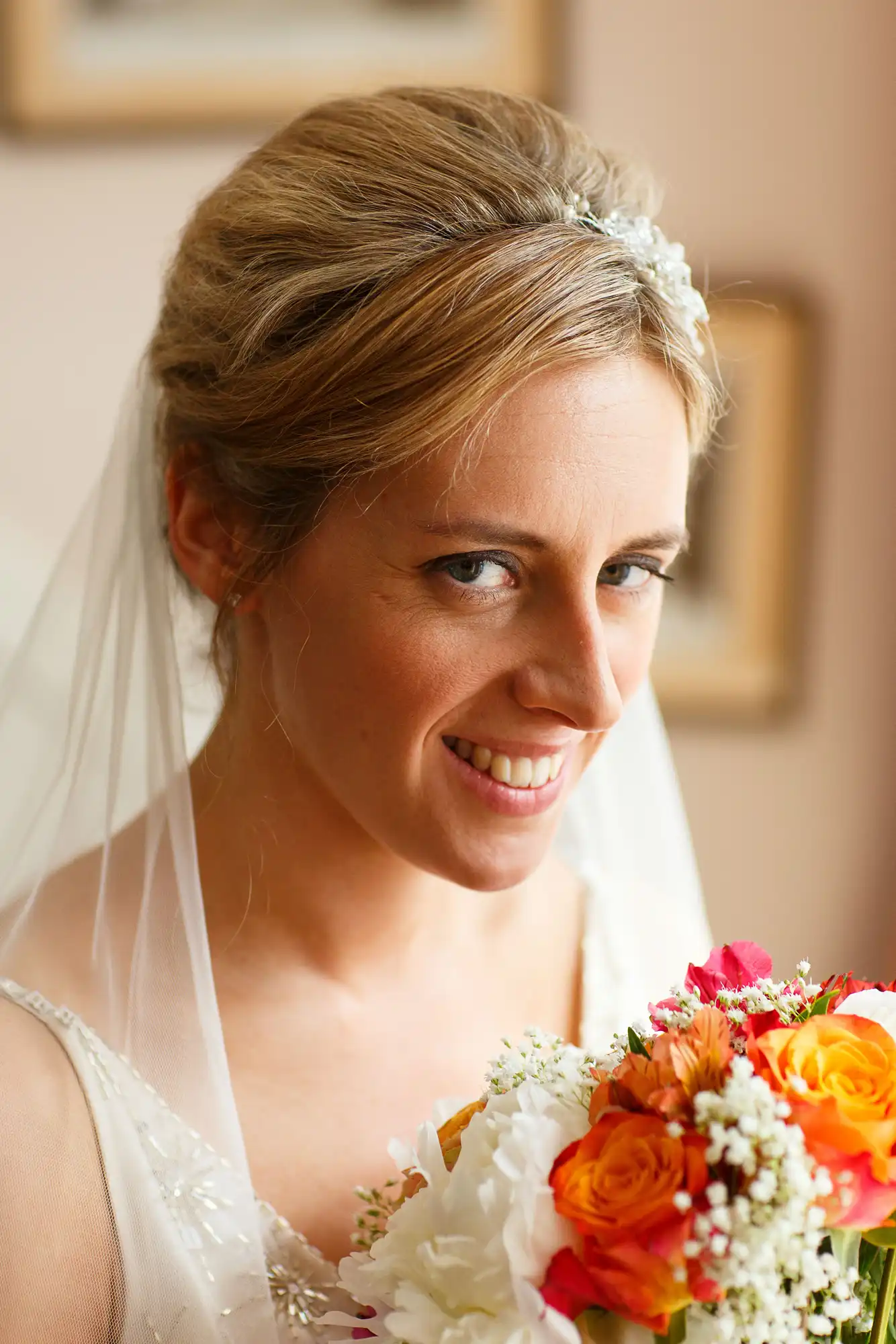 A bride smiling gently, holding a bouquet of bright orange and white flowers, wearing a white dress and a veil, with a hair accessory.