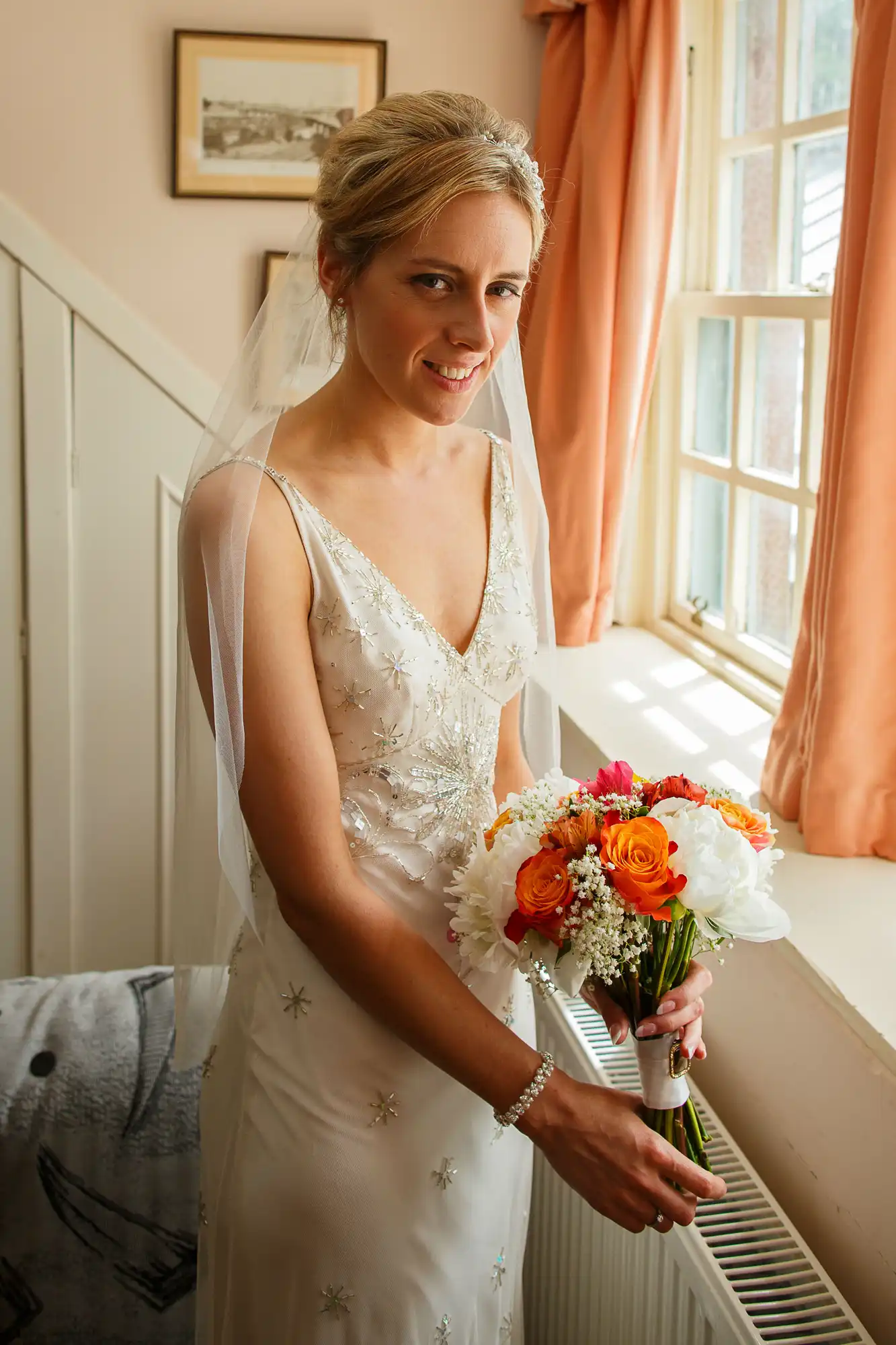 A bride in an embroidered wedding dress and veil holding a bouquet of orange and white flowers by a window with sheer curtains.