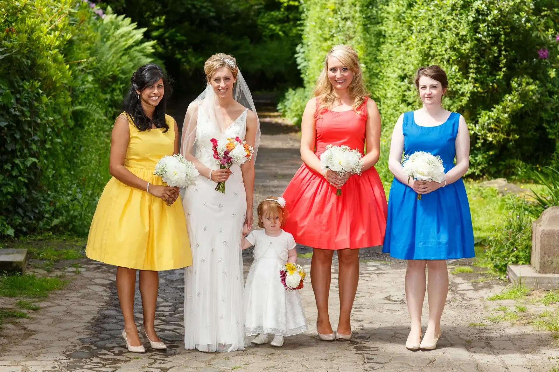 A bride in a white dress and three bridesmaids in yellow, red, and blue dresses, alongside a young flower girl, posing in a lush garden.
