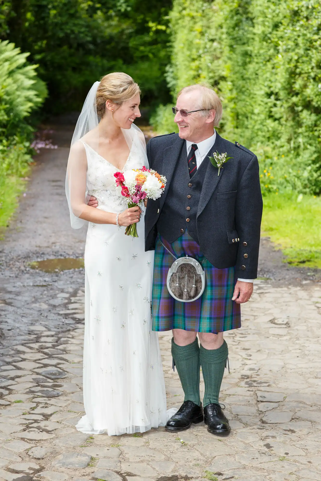 A bride in a white dress and a groom in a kilt and jacket stand arm in arm on a garden path, smiling at each other.