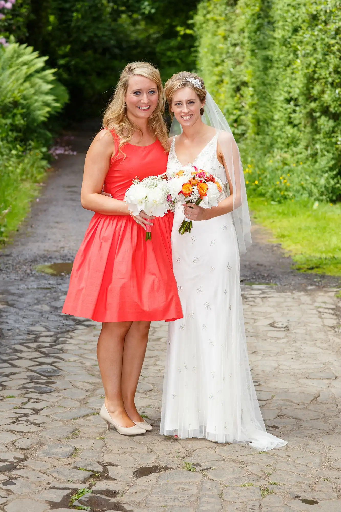 A bride in a white gown and a woman in a red dress smiling, holding bouquets on a garden path.