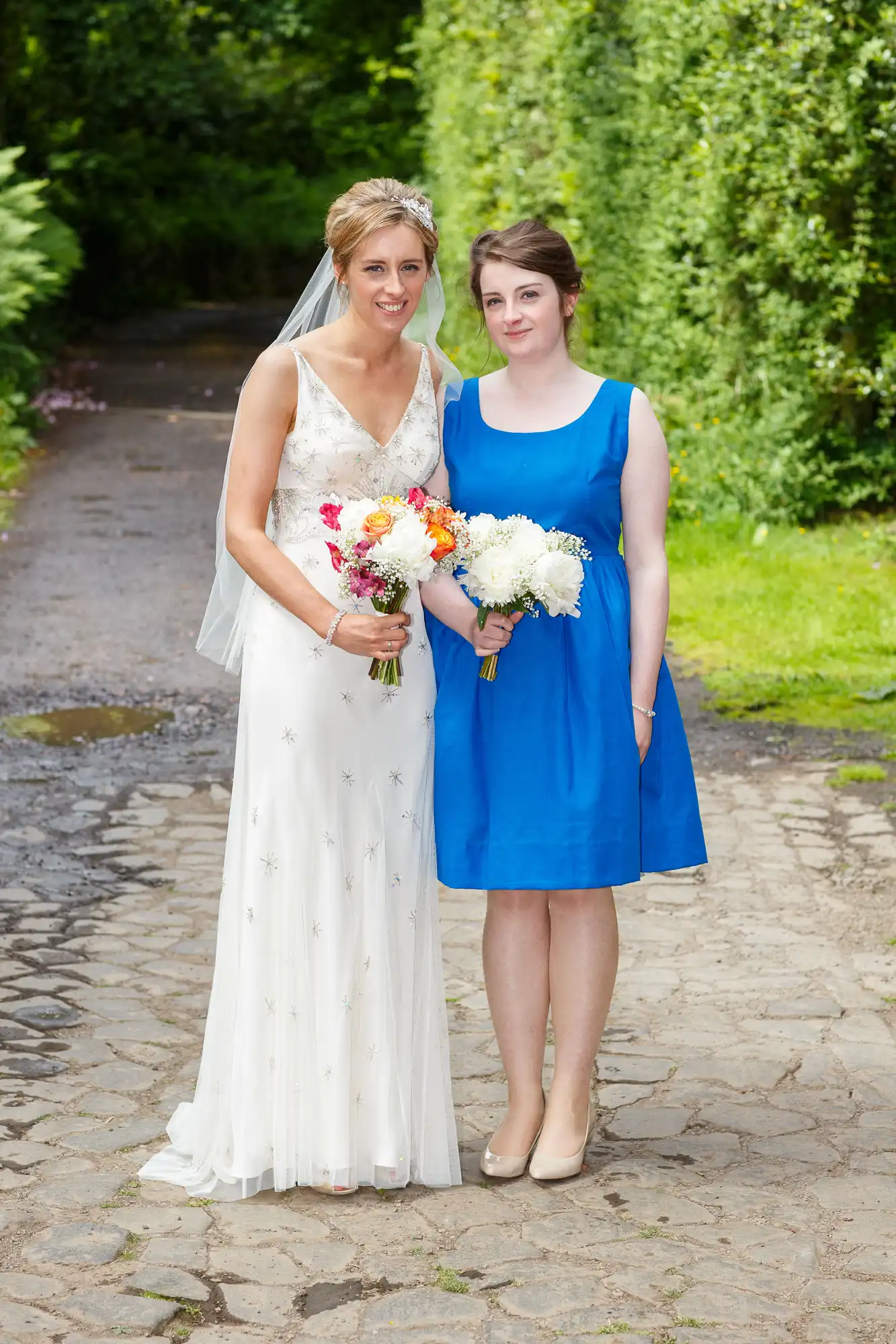 A bride in a white gown holding a bouquet stands next to a bridesmaid in a blue dress, also holding flowers, on a garden path.