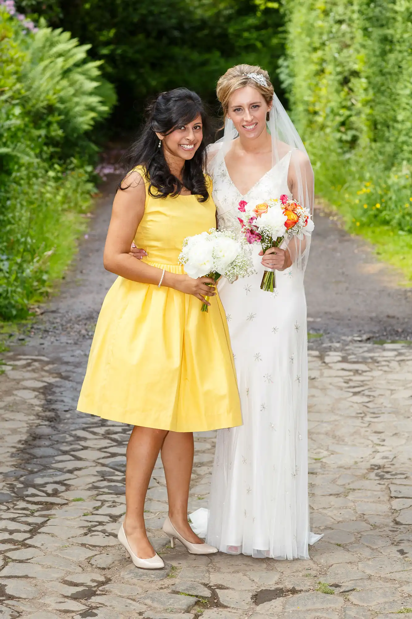 Two women, one in a yellow dress and the other in a white bridal gown, smiling and holding bouquets on a garden path.