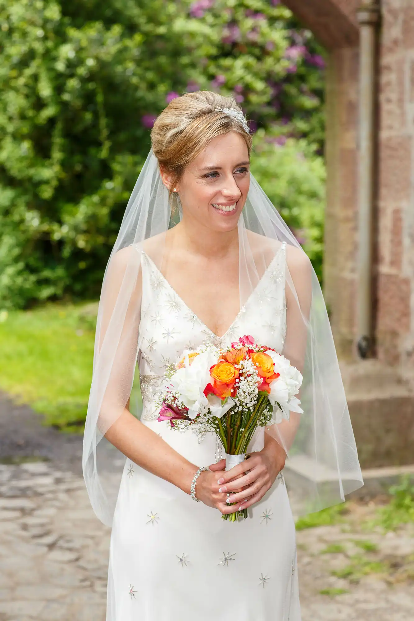 A bride in a white dress with a veil holding a bouquet of white and orange flowers, smiling outdoors with greenery in the background.