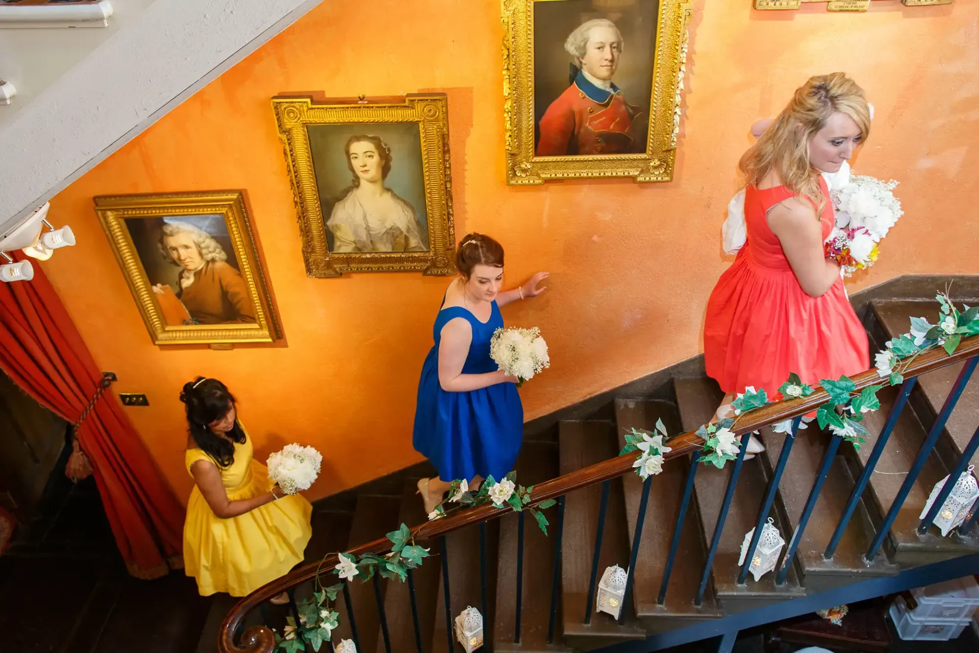 Three women in colorful dresses holding bouquets pose on a staircase adorned with greenery, surrounded by portraits on orange walls.
