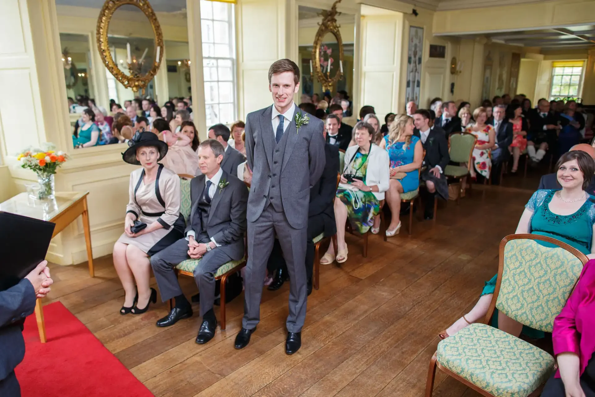A groom standing confidently at a wedding ceremony in a well-lit hall with seated guests watching him.
