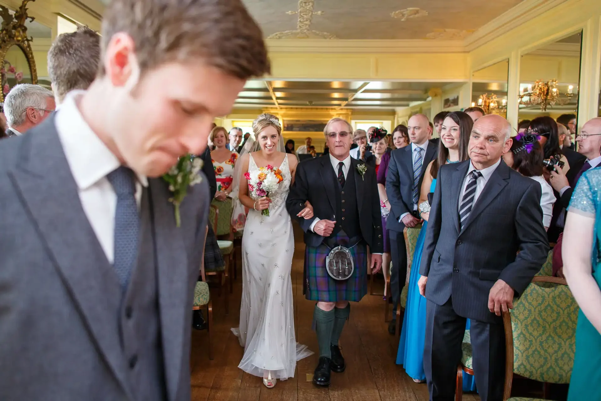 A bride walking down the aisle, accompanied by an older man in a kilt, while a groom glances downward and guests watch.