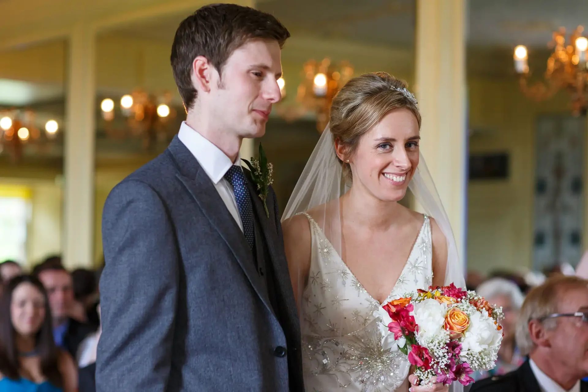 A bride and groom smiling, walking down the aisle, the bride holding a colorful bouquet, in a room filled with guests.