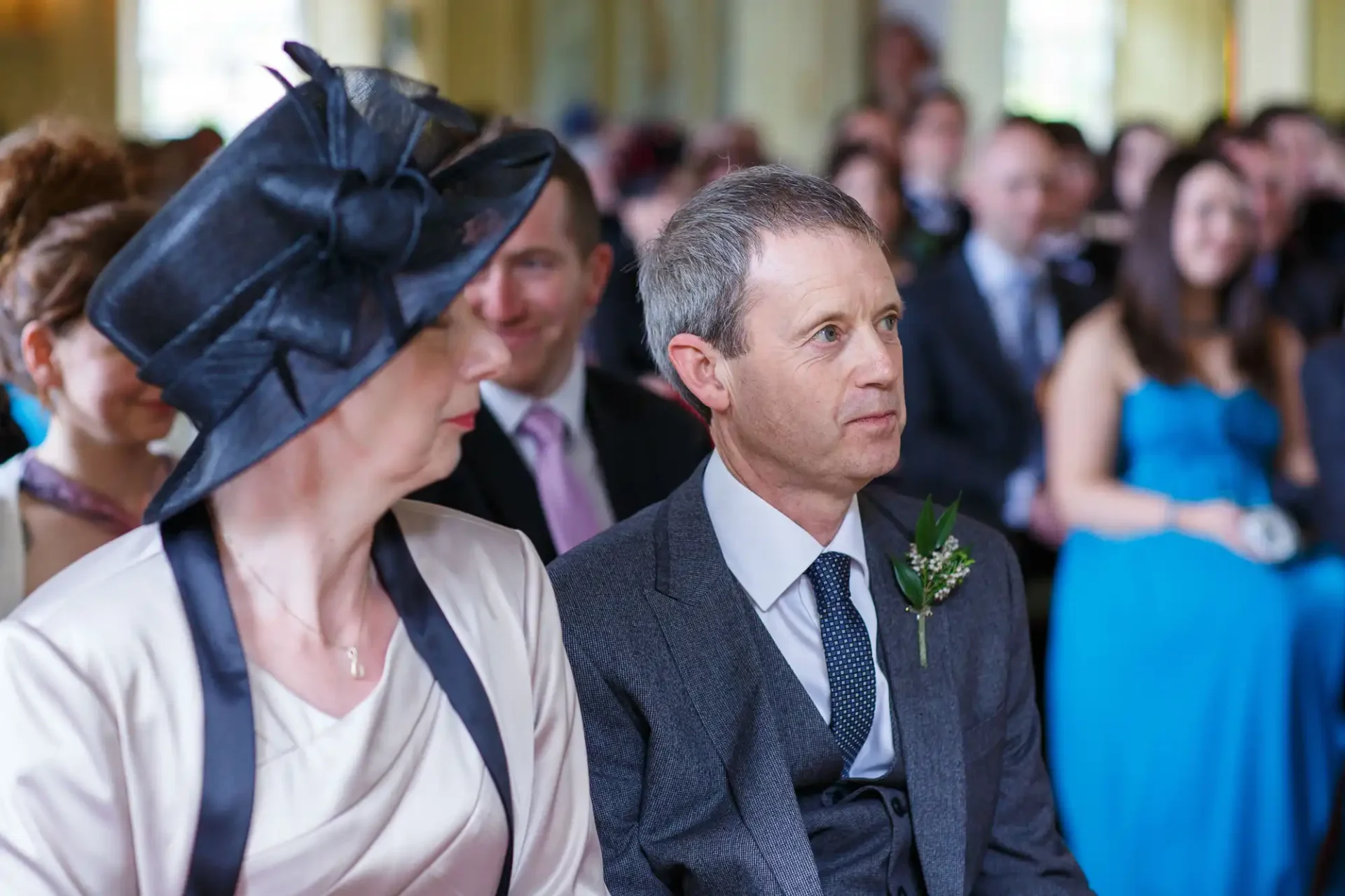 A man and a woman, dressed in formal attire, attentively watching a ceremony. the woman wears a black hat, and the man has a flower boutonniere.