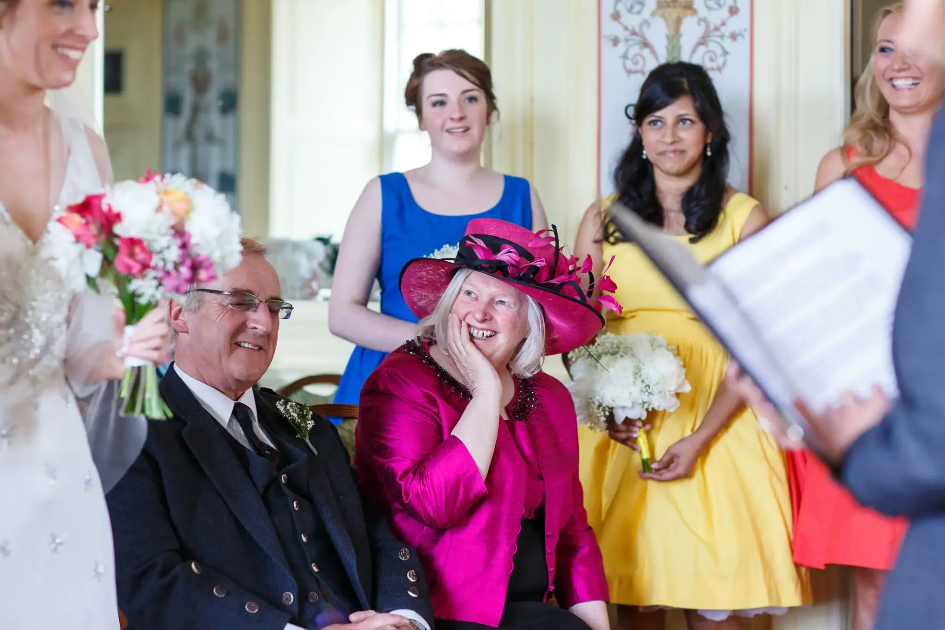 A joyful wedding scene with guests smiling and an older woman in a pink hat laughing, surrounded by people including a bride holding flowers.