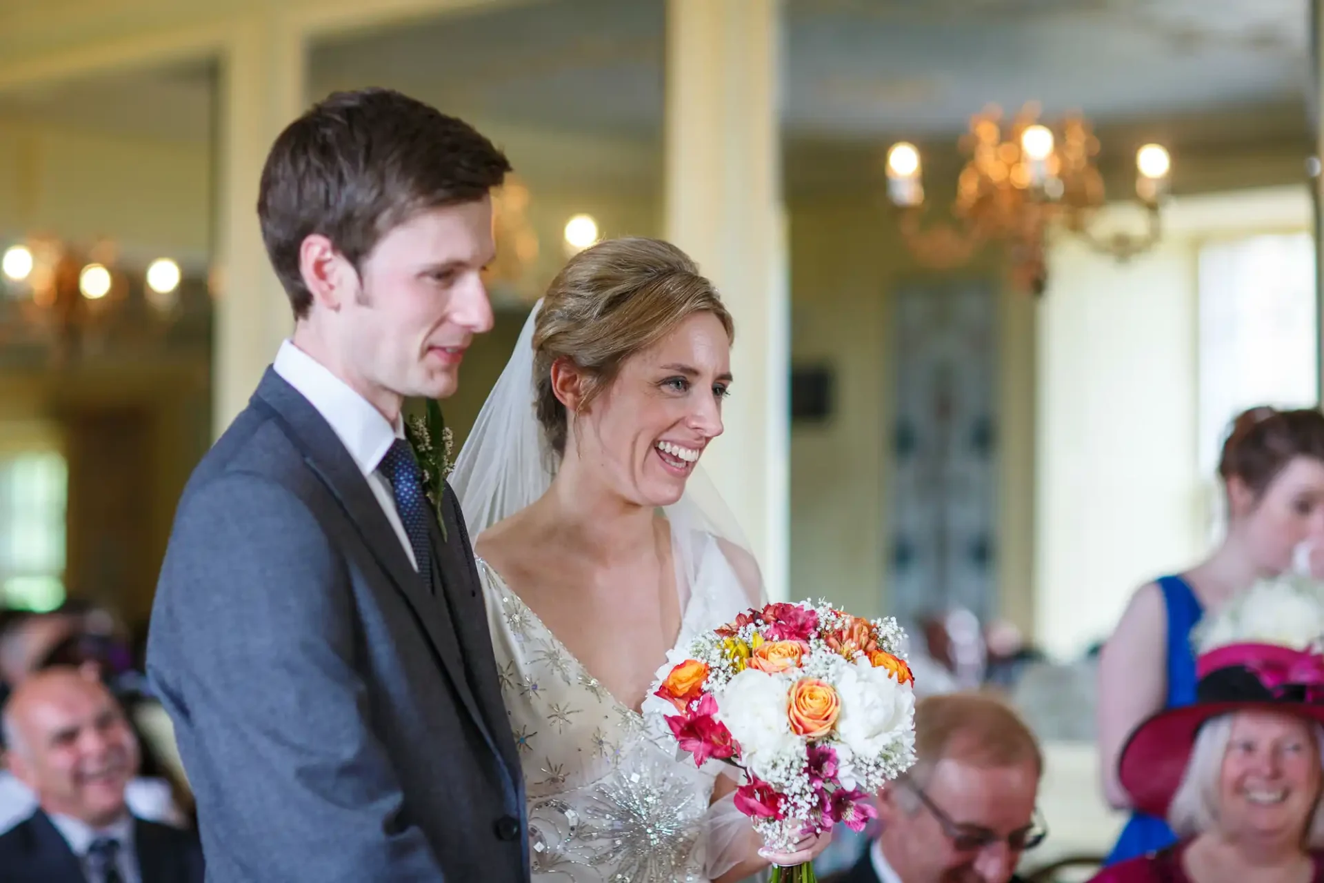 Bride holding a colorful bouquet smiling beside groom in a gray suit, with wedding guests in the background.