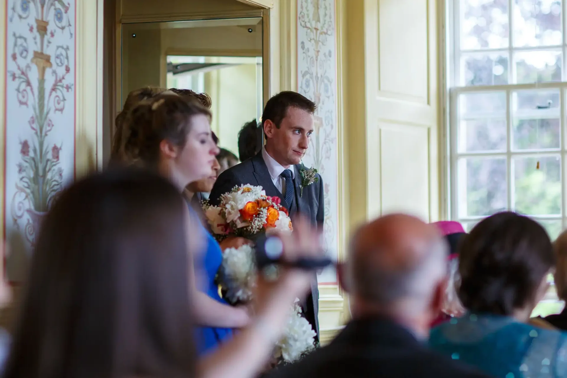 A groom seated beside a bridesmaid holding a bouquet, listening attentively during a wedding speech in a room with guests.