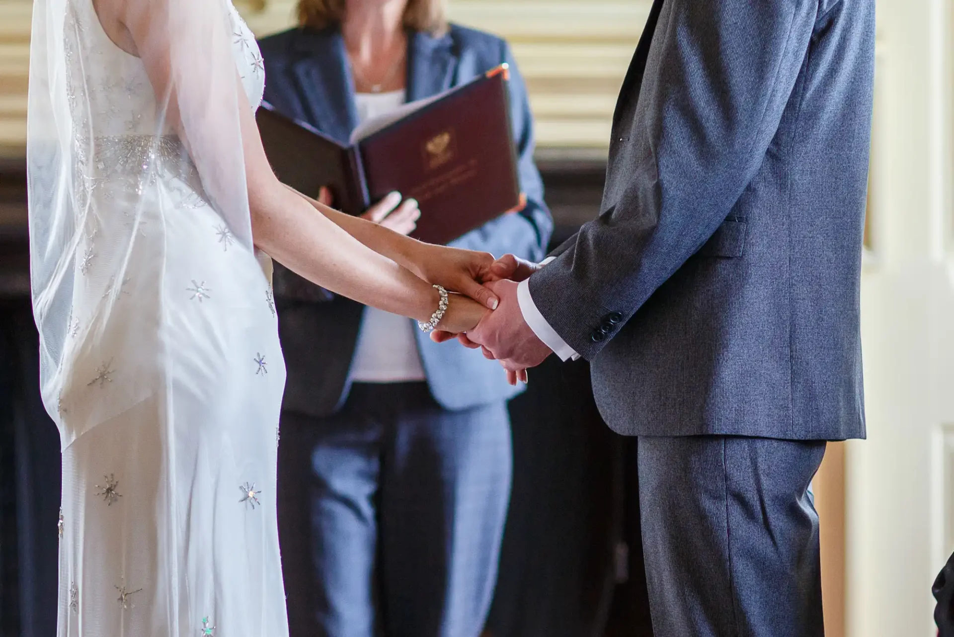 A bride in a white, embellished dress and a groom in a dark suit hold hands during a wedding ceremony officiated by a woman holding a book.