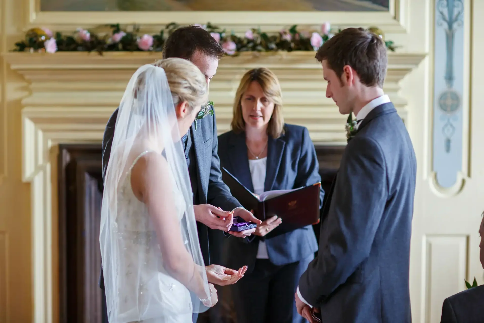 A bride and groom exchange rings during a wedding ceremony officiated by a female celebrant in an elegant room.