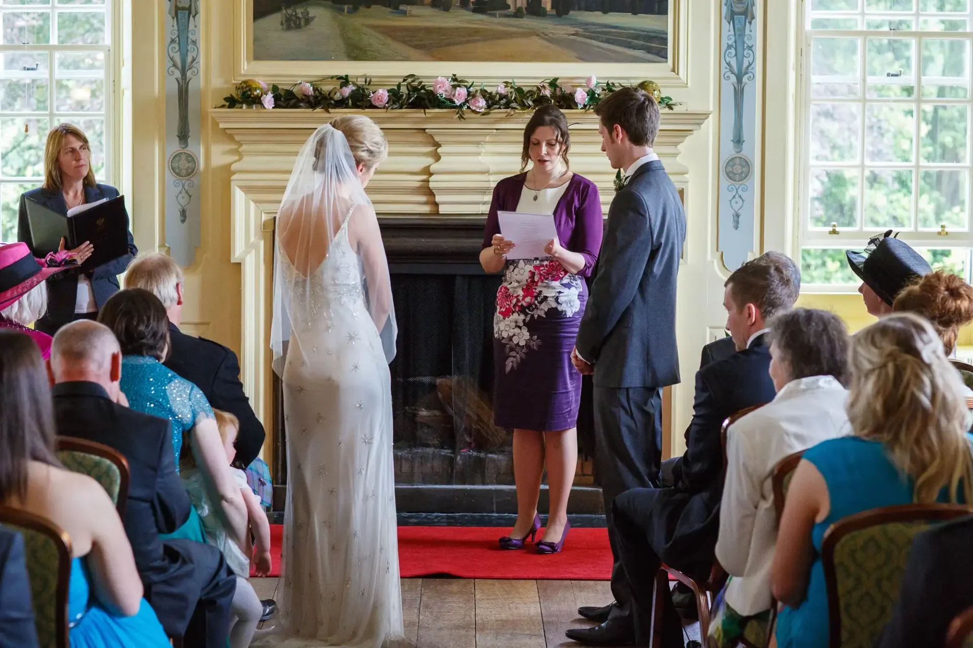 A bride and groom stand before an officiant during a wedding ceremony, with guests seated around them in a decorated room.