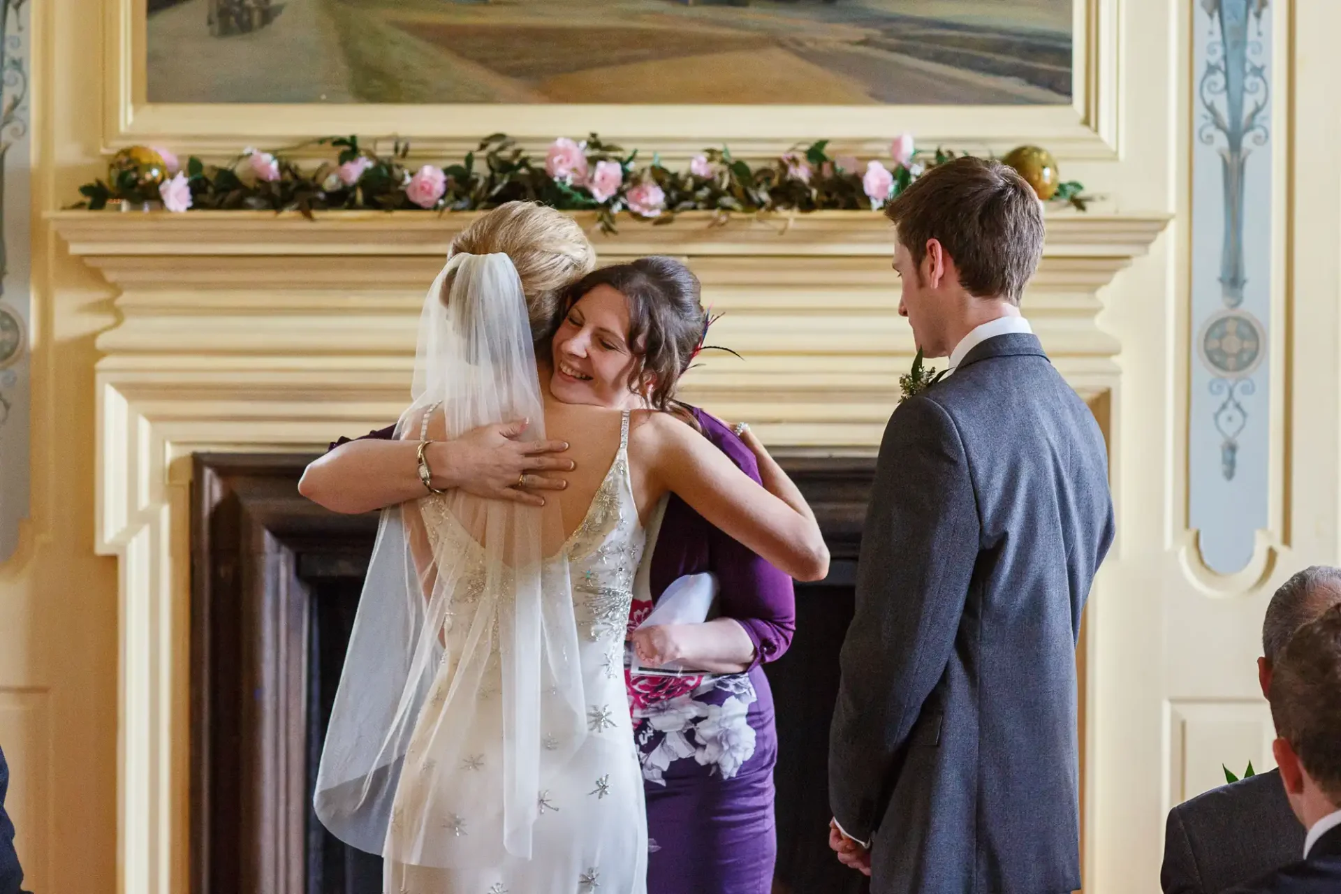A bride in a white dress and veil embraces a woman in a purple dress, while a man in a gray suit looks on, inside an elegantly decorated room.