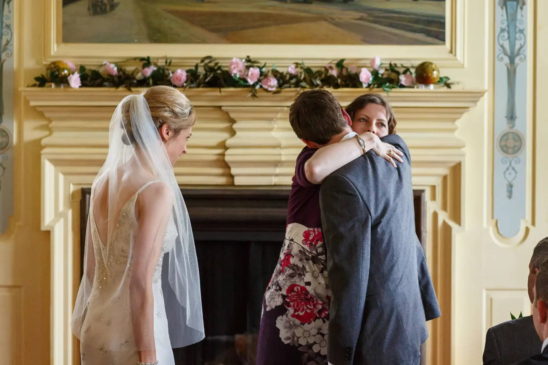 A bride watches as a wedding guest hugs another guest in front of a decorative fireplace adorned with flowers.