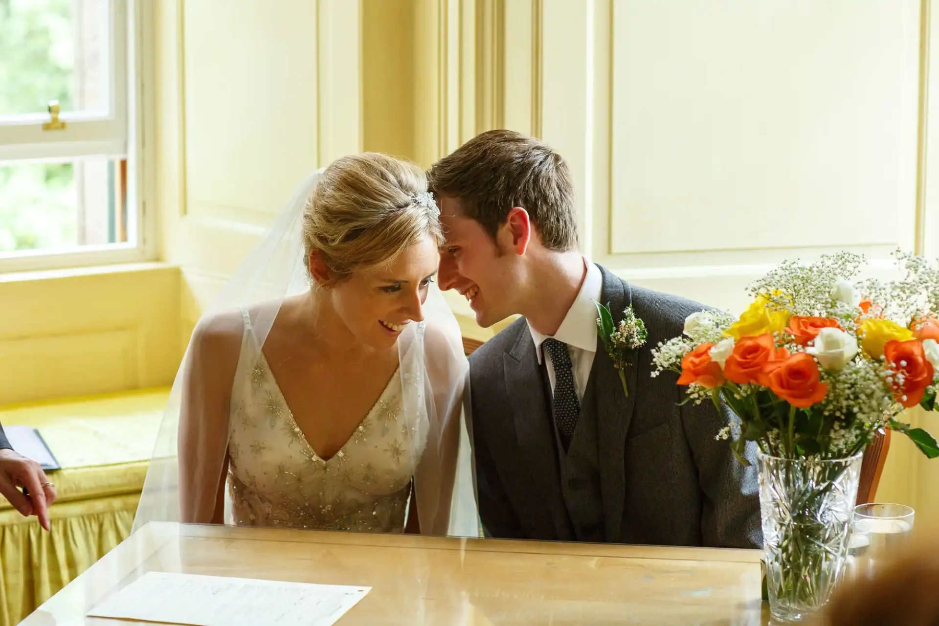 A bride and groom smiling at each other during their wedding ceremony, with a bouquet of flowers in the foreground.