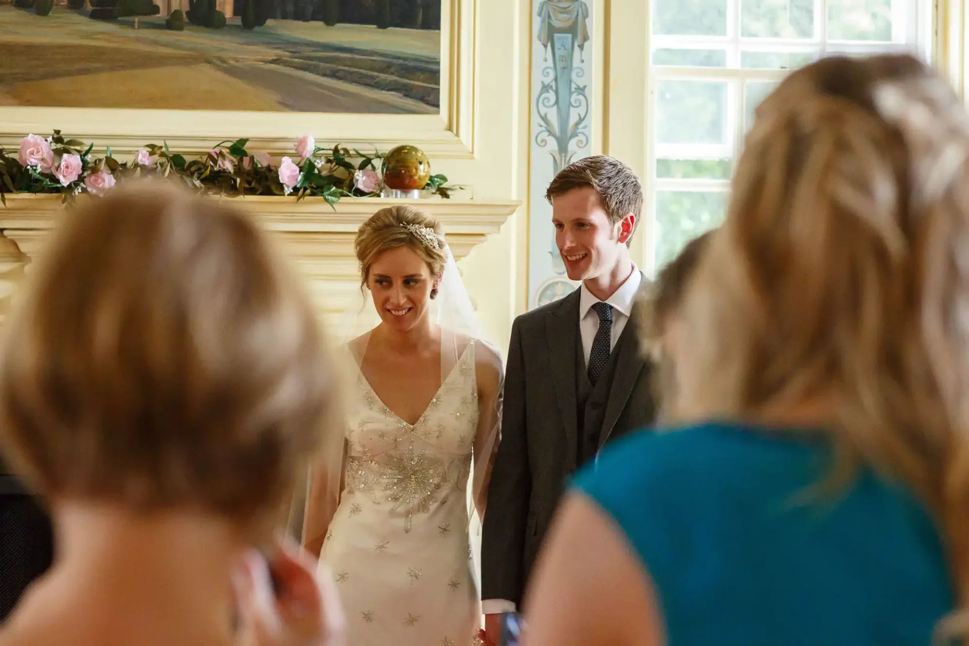 A bride and groom walking through a room smiling, surrounded by guests in an elegant interior with floral decorations.