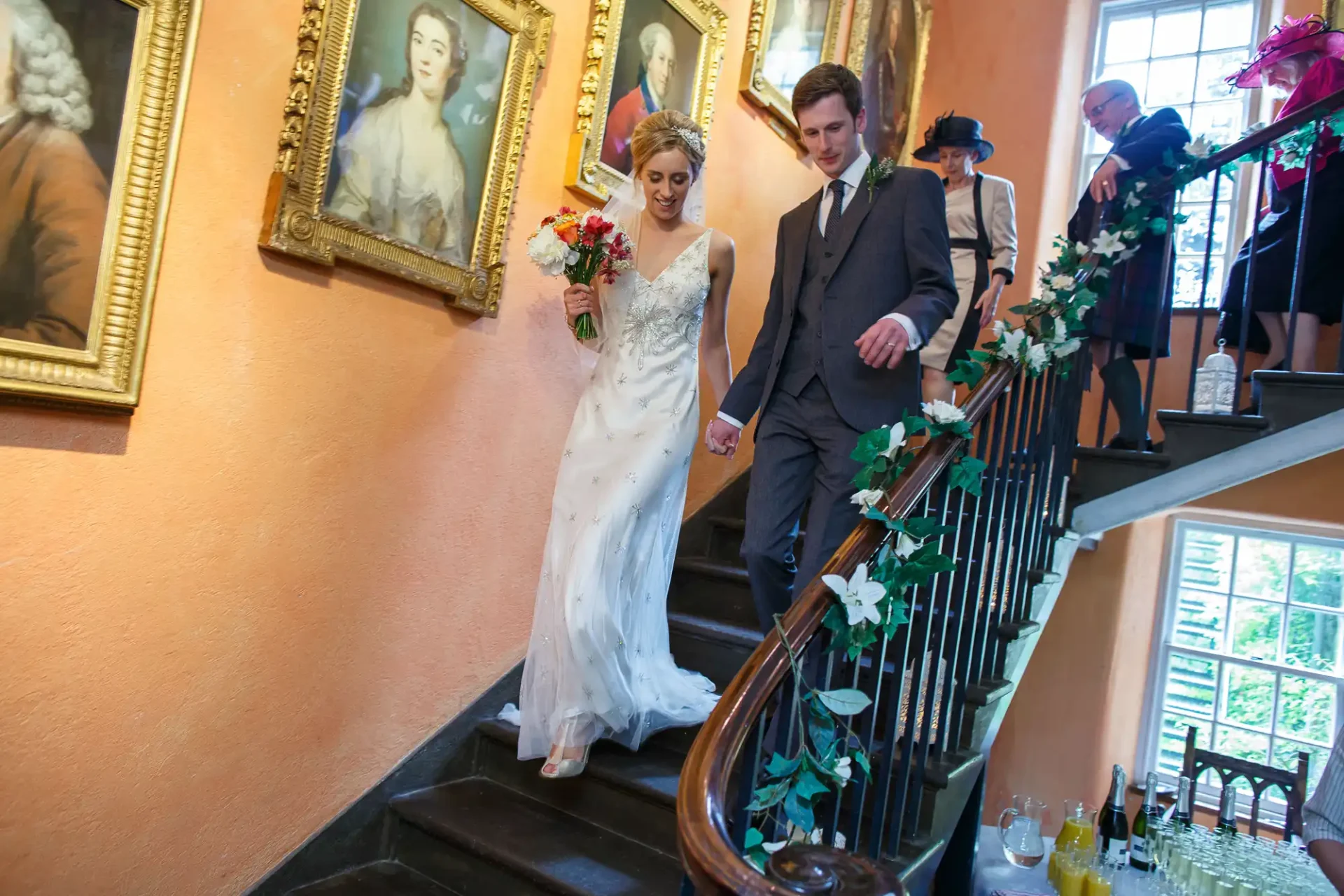 A bride and groom descend a staircase holding hands, surrounded by guests and paintings on the wall, during a wedding celebration.