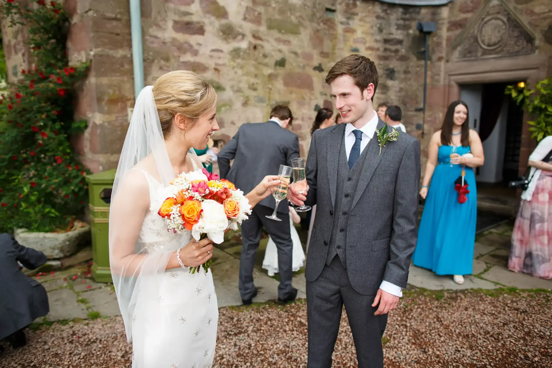 A bride and groom smiling at each other, holding glasses of wine, with guests in the background at a wedding.