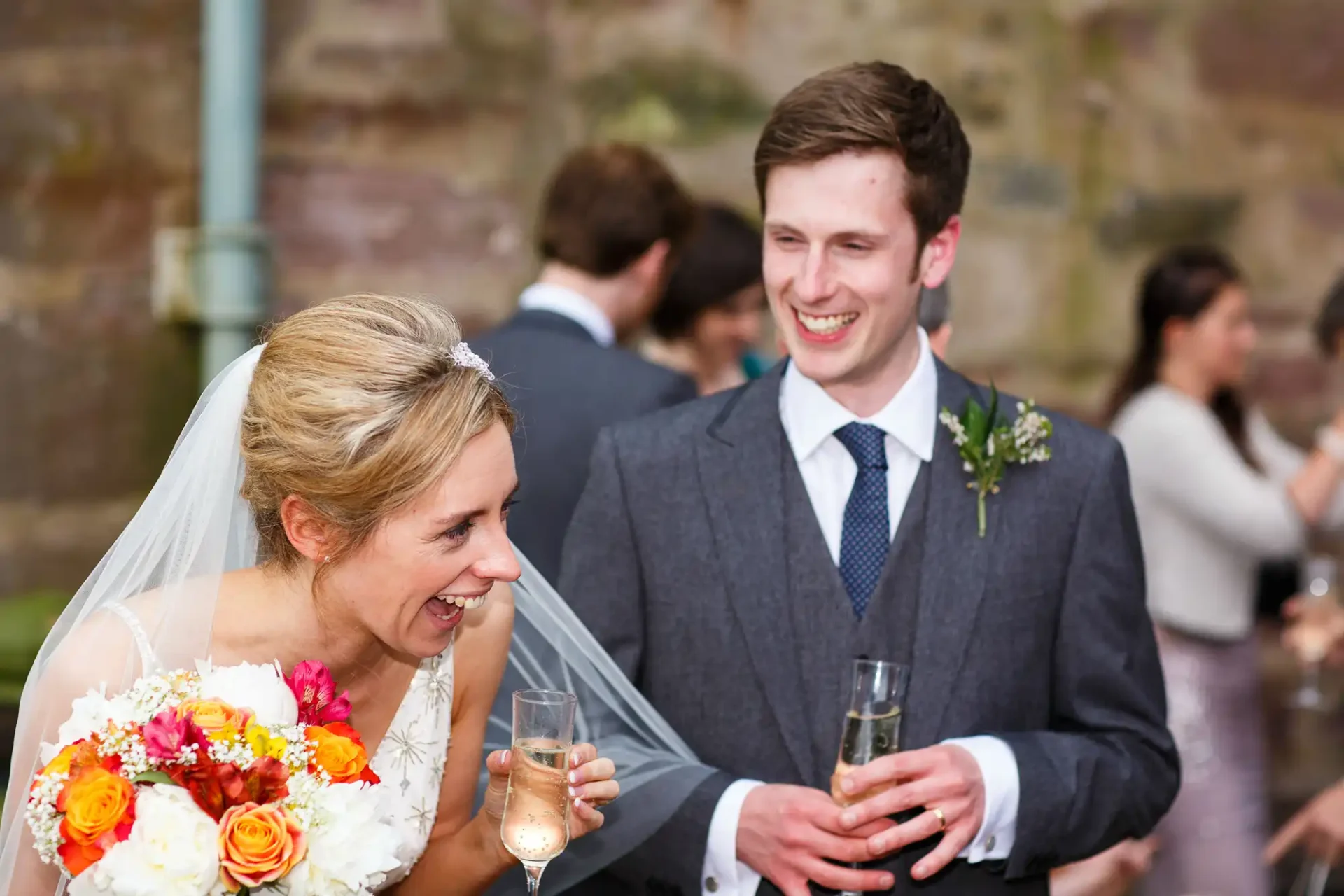 Bride and groom smiling and holding champagne glasses at a wedding reception, surrounded by guests.