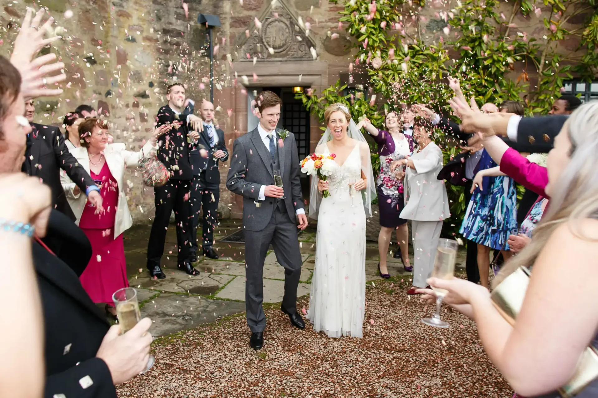 A bride and groom walk joyfully through a confetti shower, surrounded by cheering guests at an outdoor wedding venue.