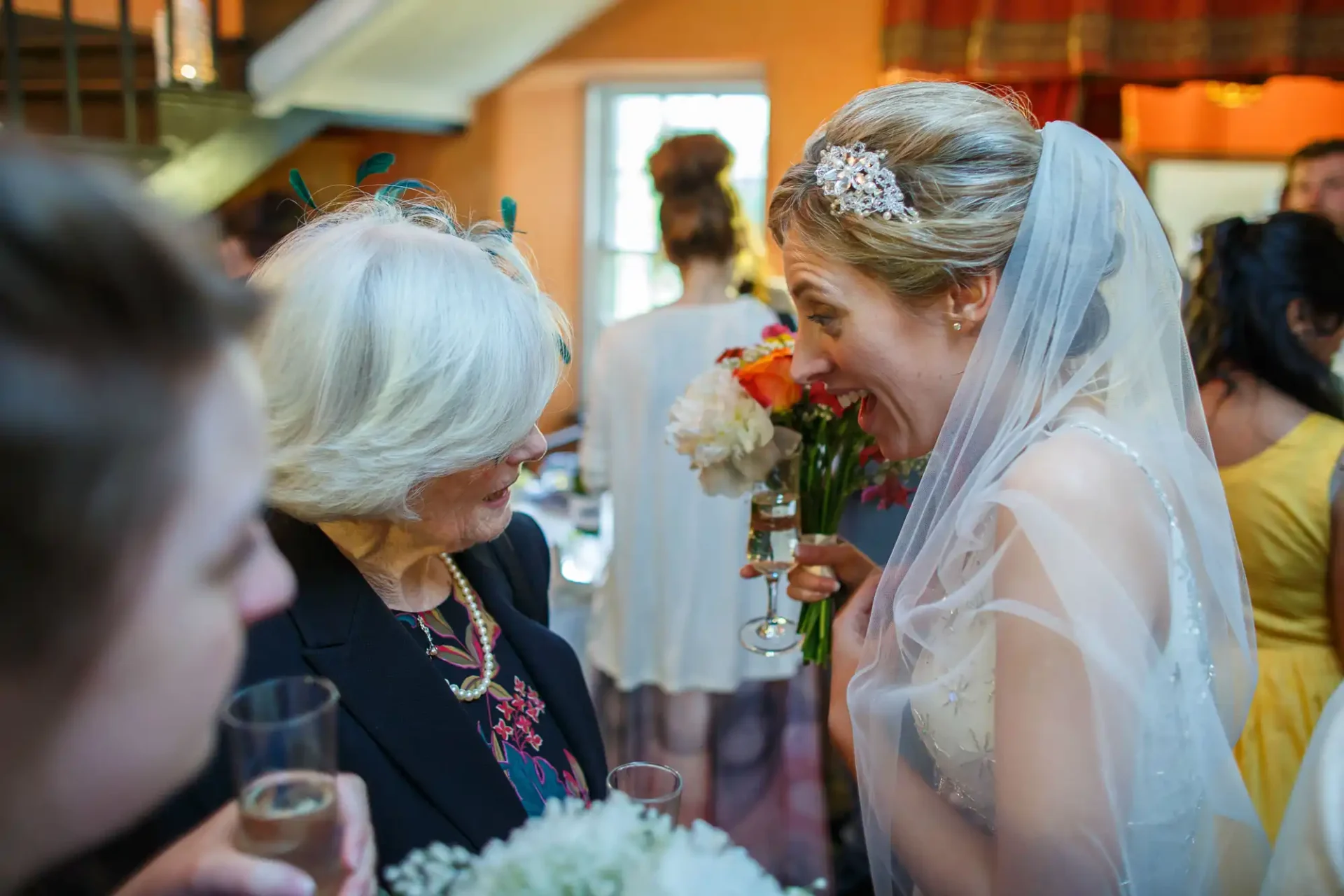 A bride in a veil smiles broadly while talking to an elderly woman at a wedding reception, each holding a glass.