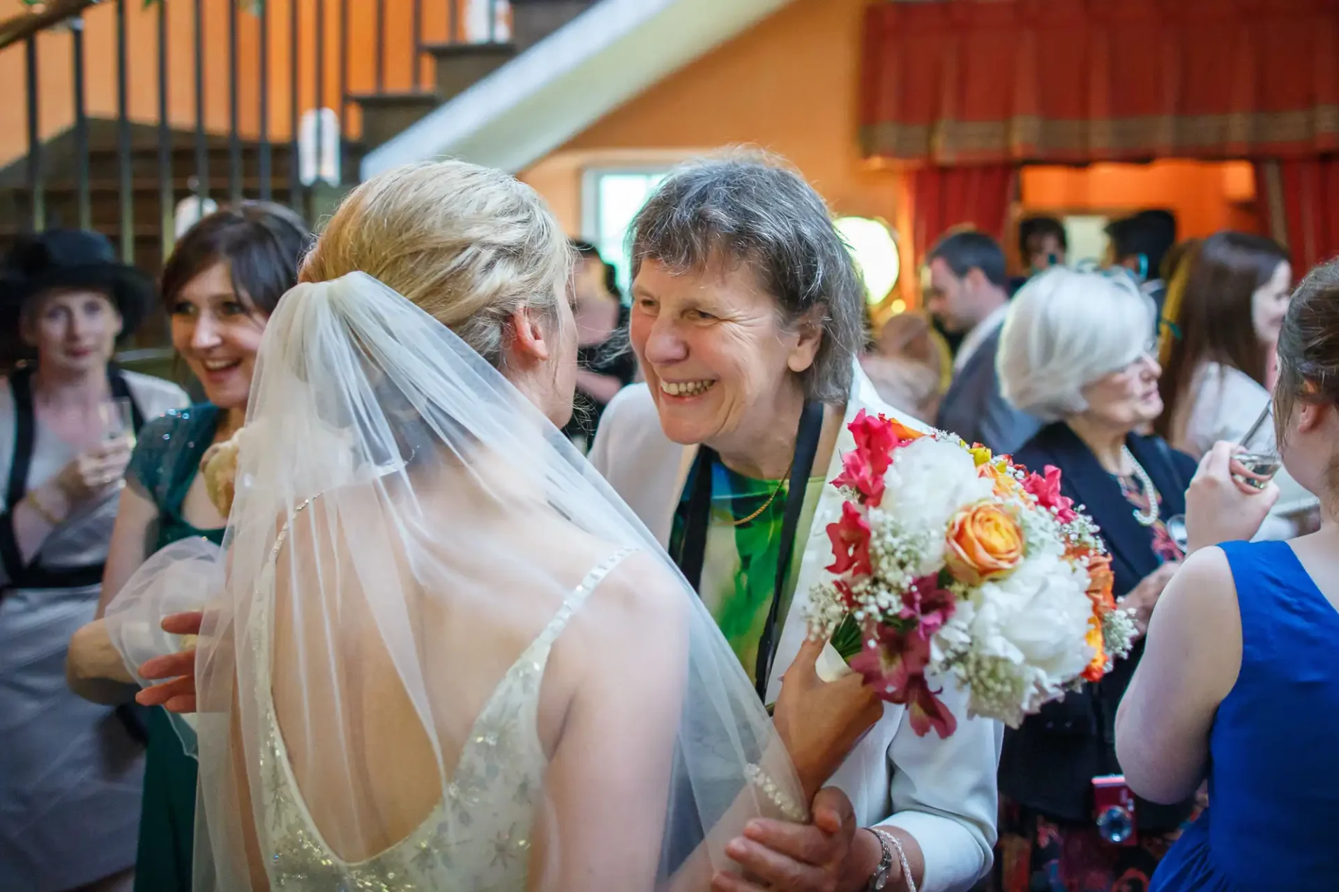A bride conversing with an older woman holding flowers at a wedding, surrounded by guests.