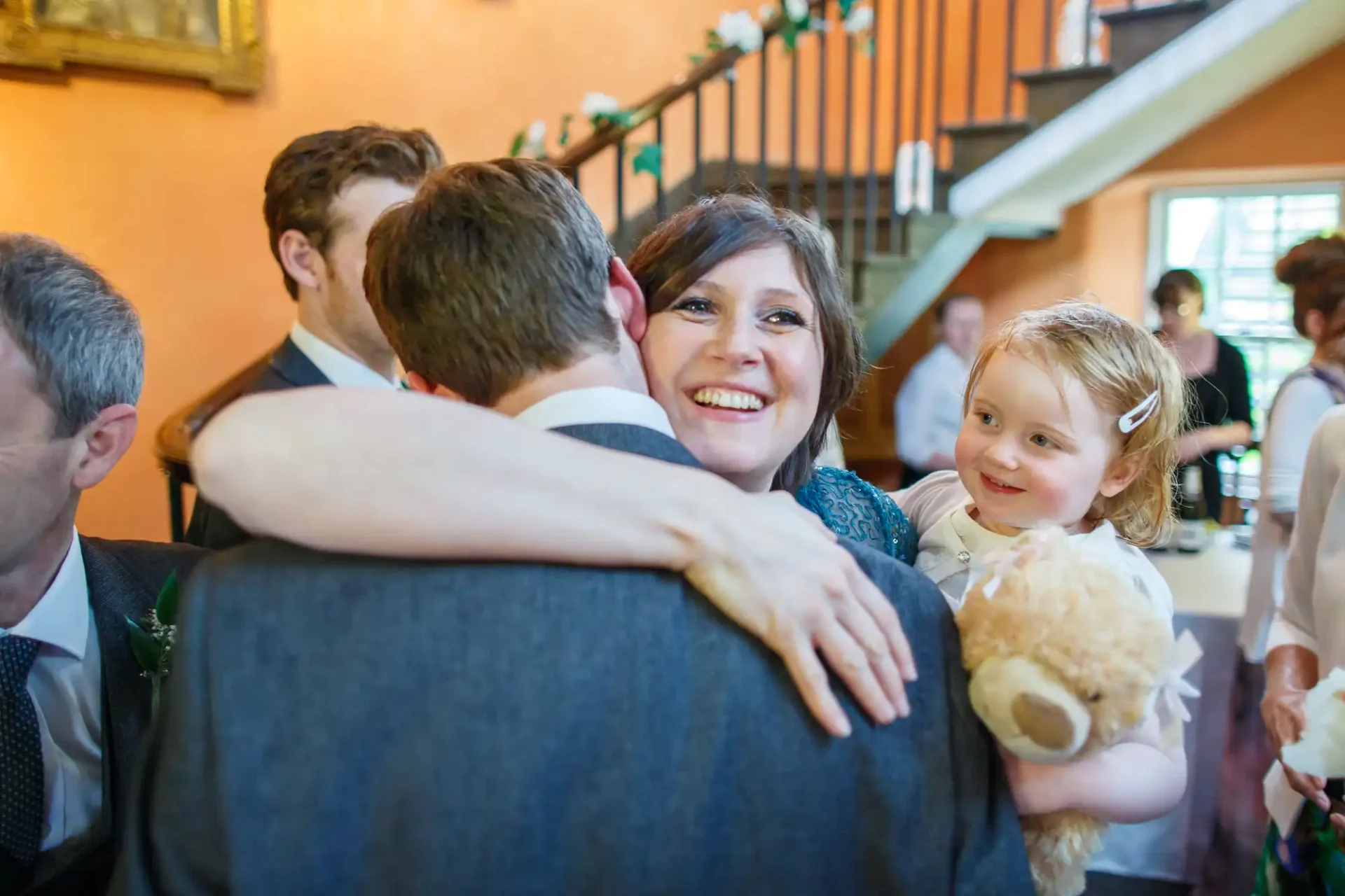 A joyful woman with short brown hair hugs a man at a wedding, holding a little girl with a teddy bear; other guests appear in the background.