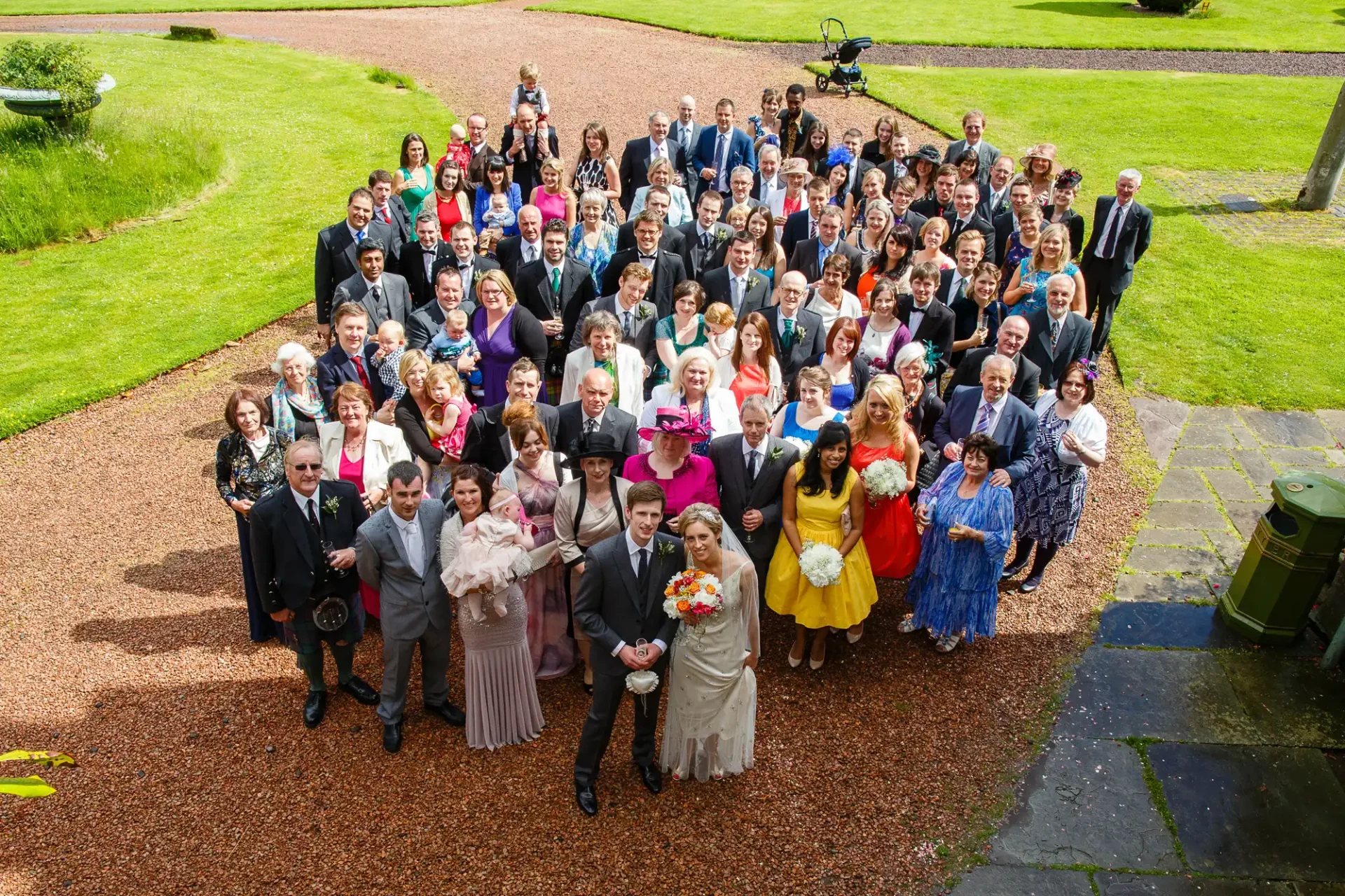 A large group of wedding guests, dressed formally, posing for a photo in a heart-shaped arrangement on a gravel courtyard with green lawns in the background.