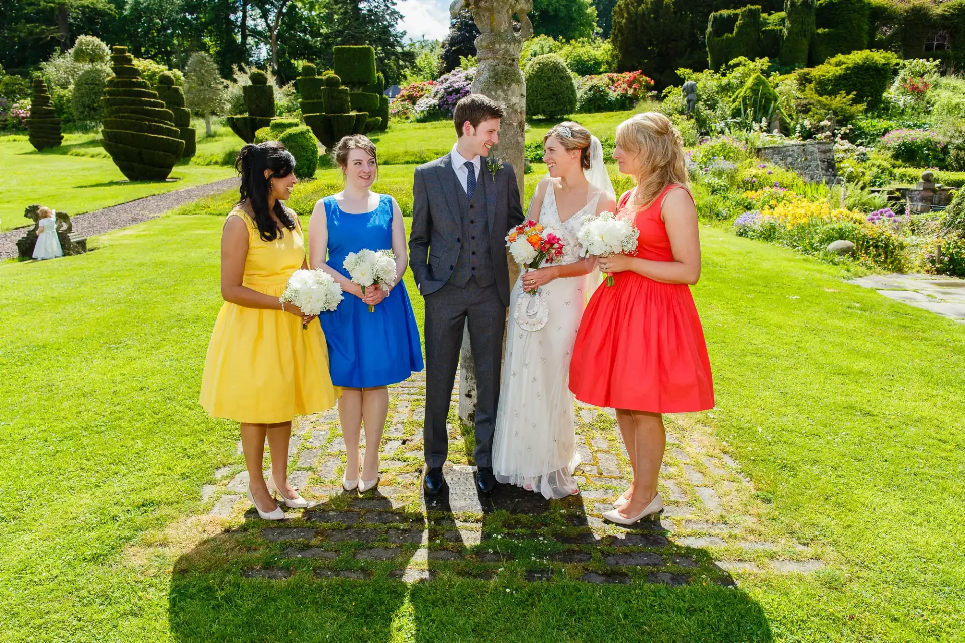 A wedding couple and three bridesmaids, dressed in colorful dresses, standing in a lush garden under bright sunlight.