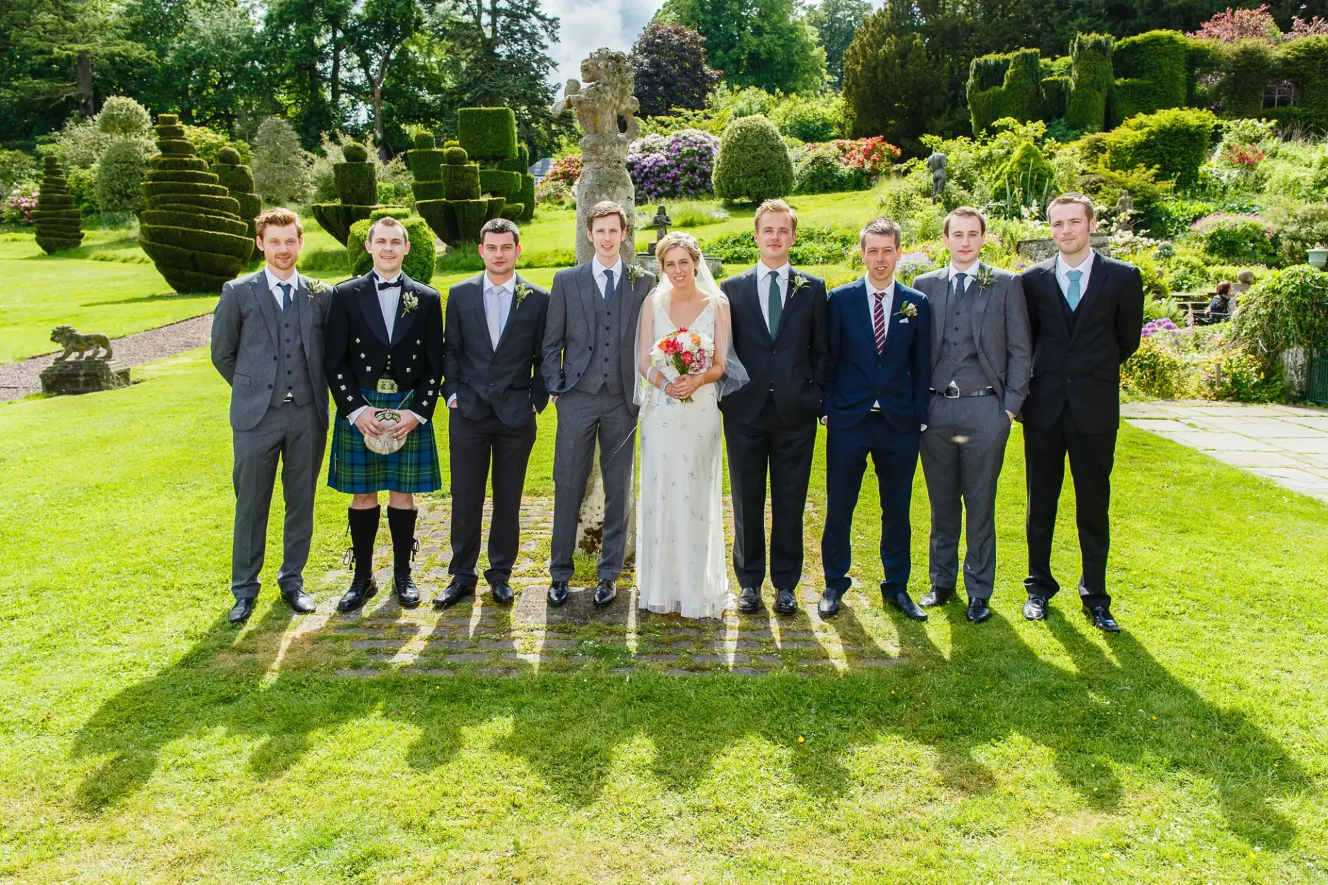 A bride and groom with six groomsmen posing in a formal garden; one groomsman wearing traditional scottish attire.