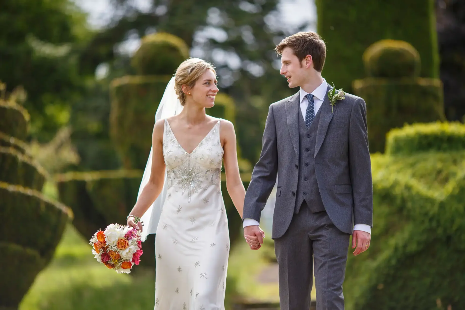 A bride and groom holding hands and smiling at each other while walking in a garden, the bride carrying a bouquet.