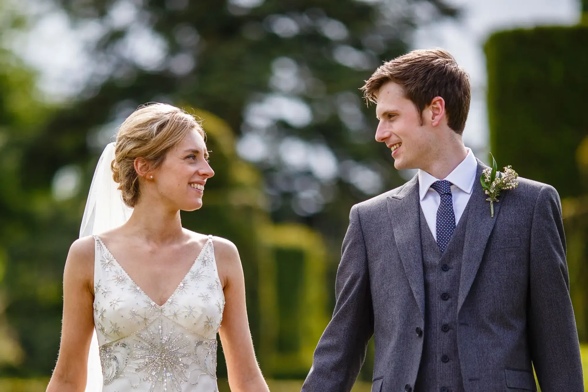A bride and groom smiling at each other outdoors on a sunny day, both dressed in wedding attire.