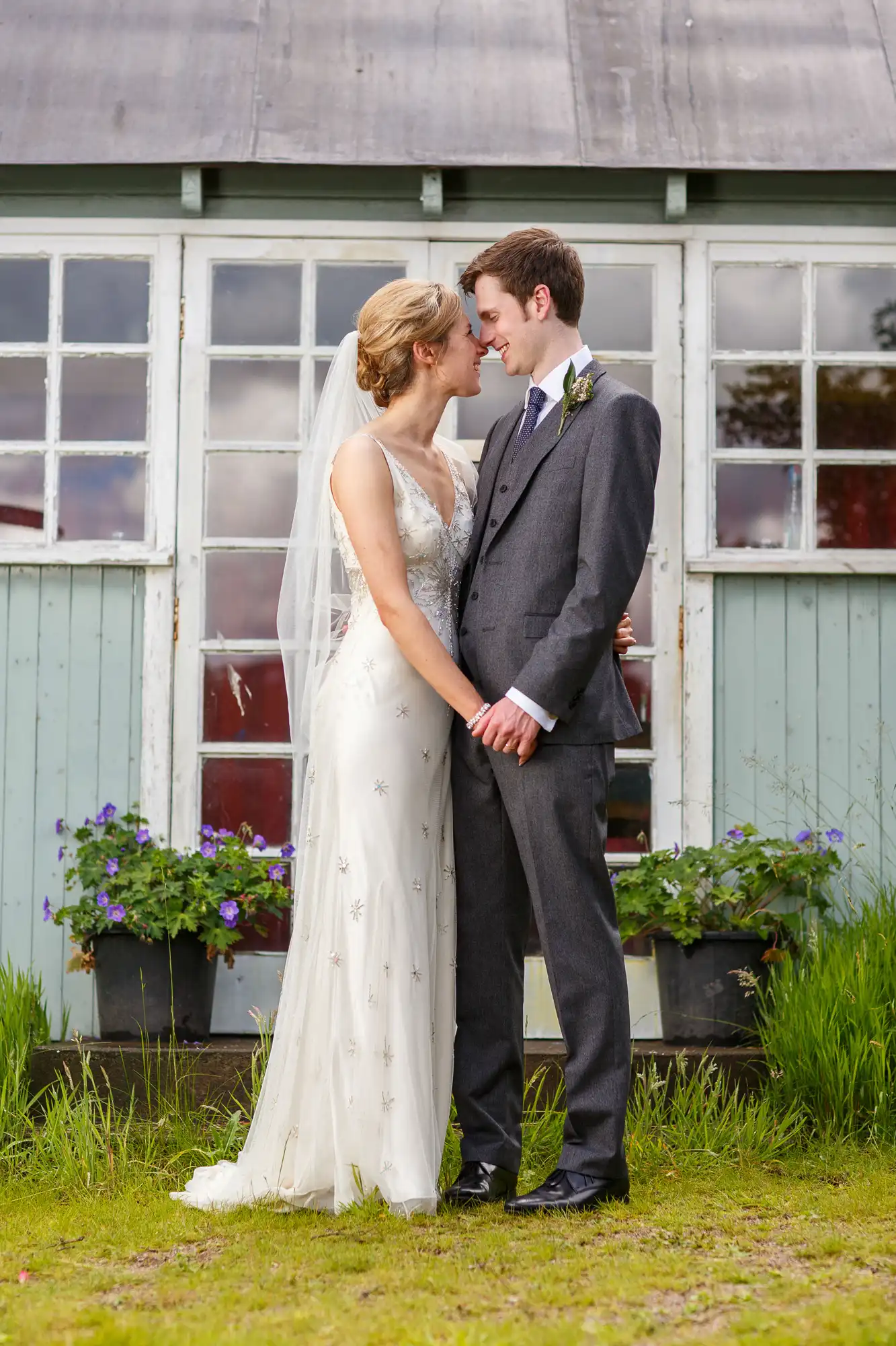 A bride and groom holding hands and touching foreheads gently in front of a rustic building with large windows and surrounded by greenery.